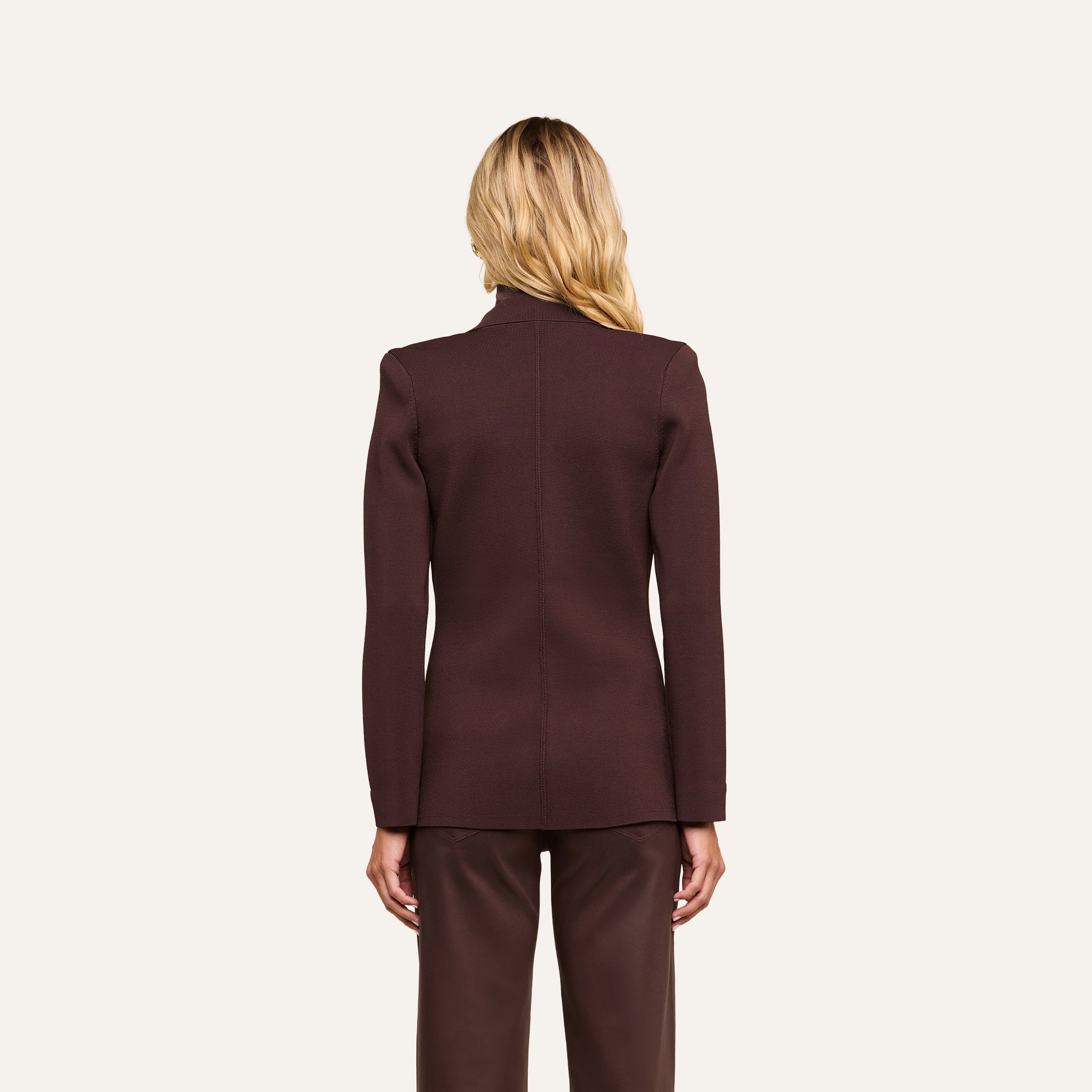 A person with long blonde hair is standing facing away, wearing the Amelia Knit Blazer and matching pants in dark brown, against a plain light background.