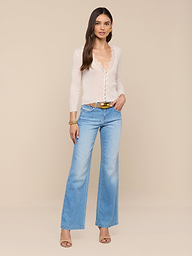 A woman stands against a white background, wearing the Eluna Cardigan Top—a sheer, featherweight knit long-sleeve—paired with light blue flared jeans, a belt, gold bracelet, and heeled sandals. She has long hair and one hand in her pocket.