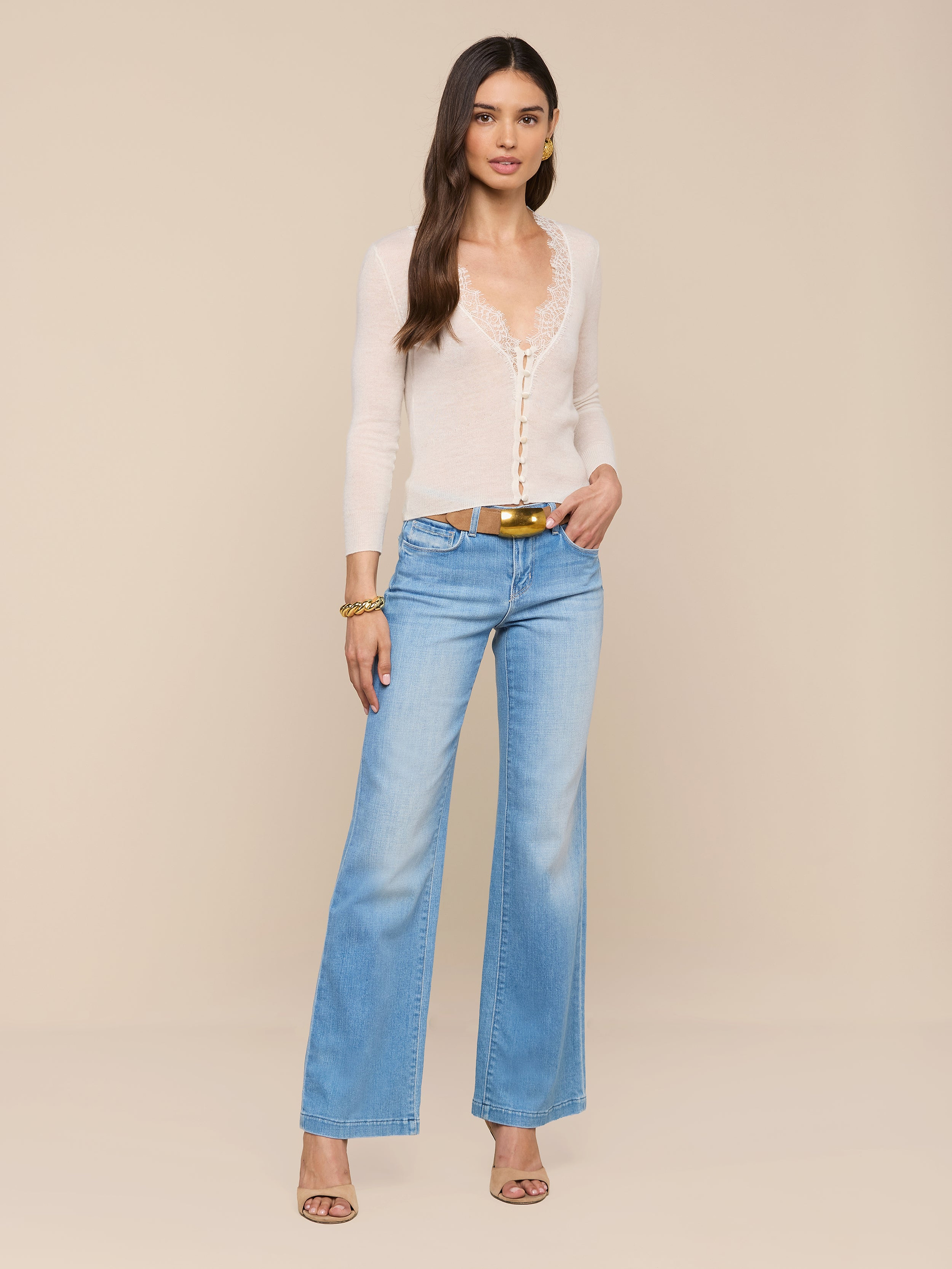 A woman stands against a white background, wearing the Eluna Cardigan Top—a sheer, featherweight knit long-sleeve—paired with light blue flared jeans, a belt, gold bracelet, and heeled sandals. She has long hair and one hand in her pocket.