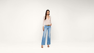 A woman wearing a sheer white top, light blue wide-leg jeans, and heels stands against a plain white background, looking at the camera with one hand in her pocket.