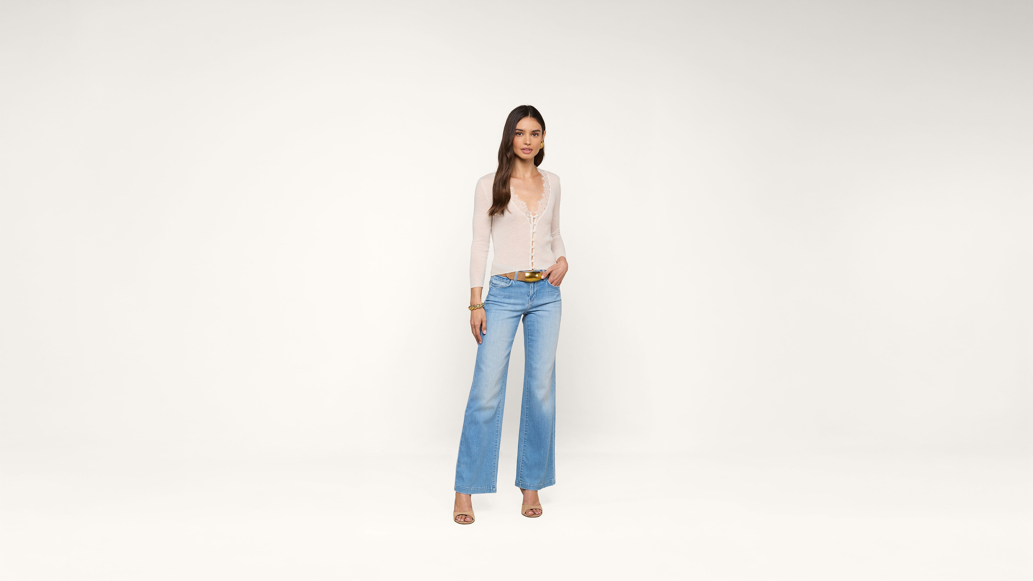 A woman wearing a sheer white top, light blue wide-leg jeans, and heels stands against a plain white background, looking at the camera with one hand in her pocket.