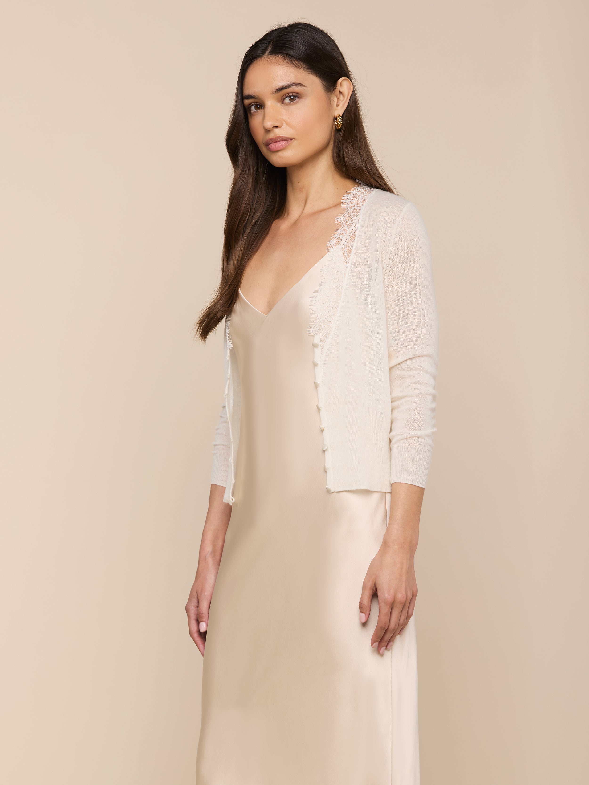 Wearing the Eluna Cardigan Top layered over a delicate, silky slip dress, a woman with long, straight hair and a neutral expression stands against a plain, light background, gazing slightly to the viewer’s left.