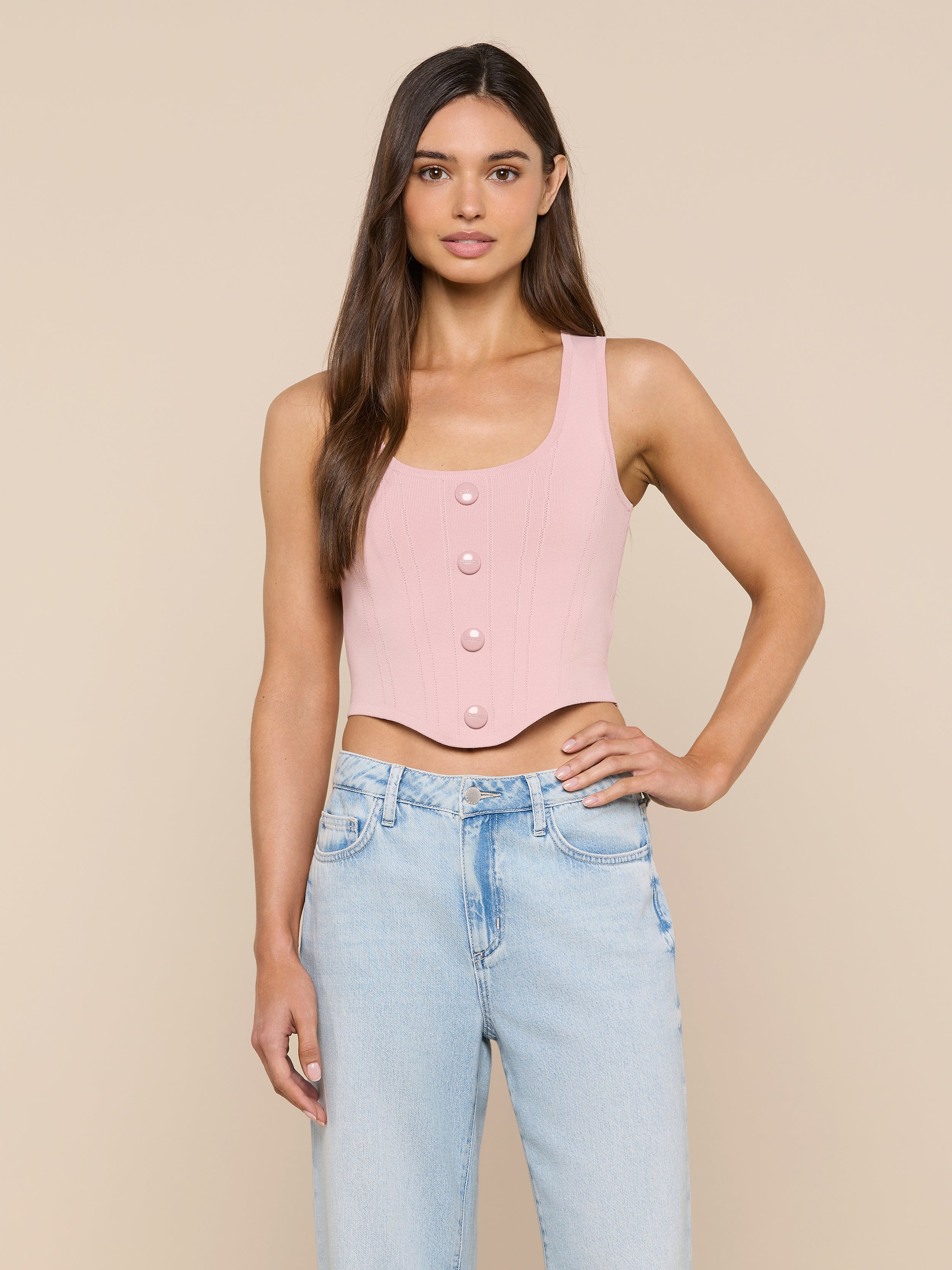 A woman stands against a plain background wearing the Dove Knit Corset Top, a light pink, sleeveless, fitted crop top with a curved hemline and button-front closure, paired with high-waisted light blue jeans. She has long straight brown hair and a neutral expression.