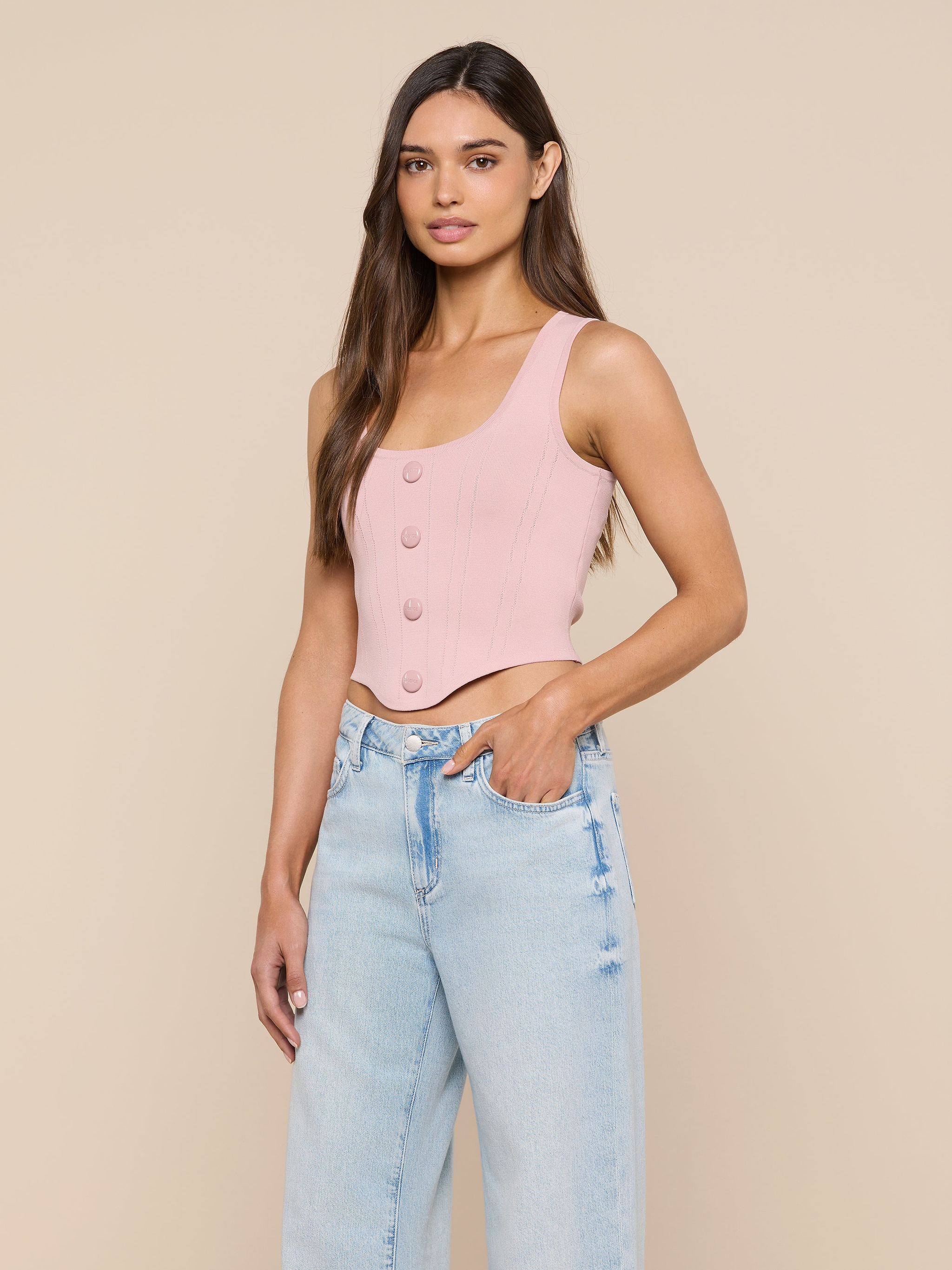 A woman wearing the Dove Knit Corset Top in light pink—a sleeveless, buttoned crop top with a curved hemline—paired with light blue high-waisted jeans, stands with one hand in her pocket against a plain off-white background.