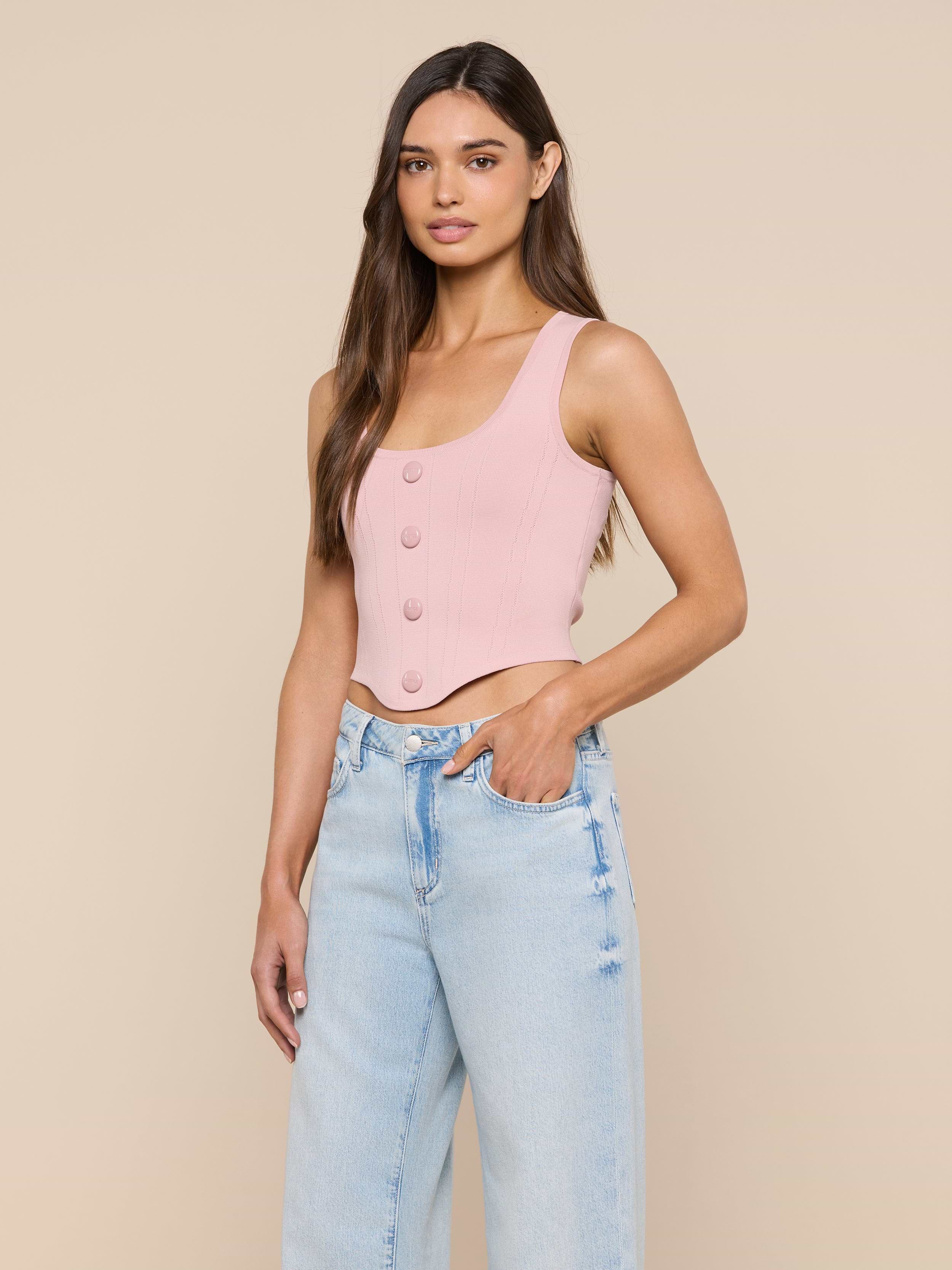 A woman wearing the Dove Knit Corset Top in light pink—a sleeveless, buttoned crop top with a curved hemline—paired with light blue high-waisted jeans, stands with one hand in her pocket against a plain off-white background.