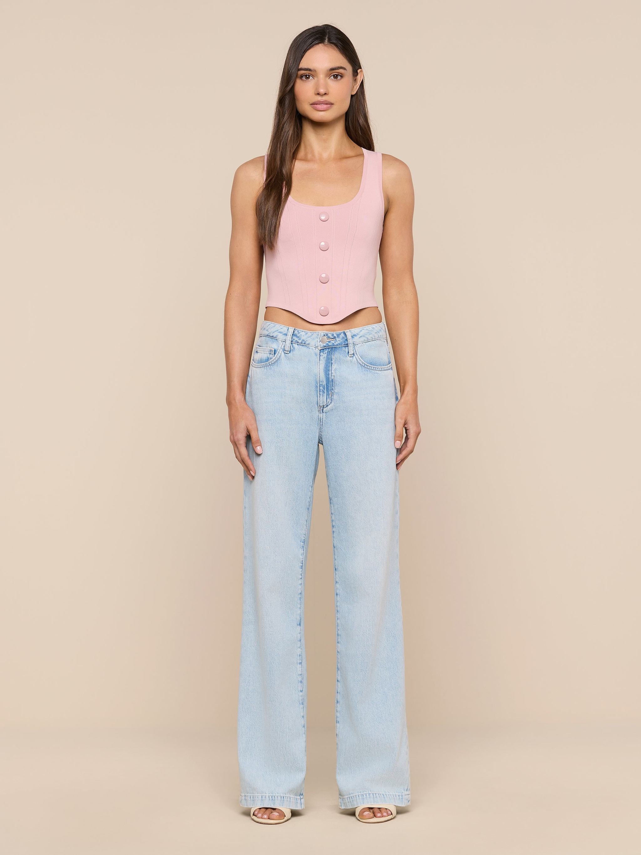 A woman stands facing forward, wearing the Dove Knit Corset Top in pink and light blue wide-leg jeans with a visible waistband detail. The background is plain and white.