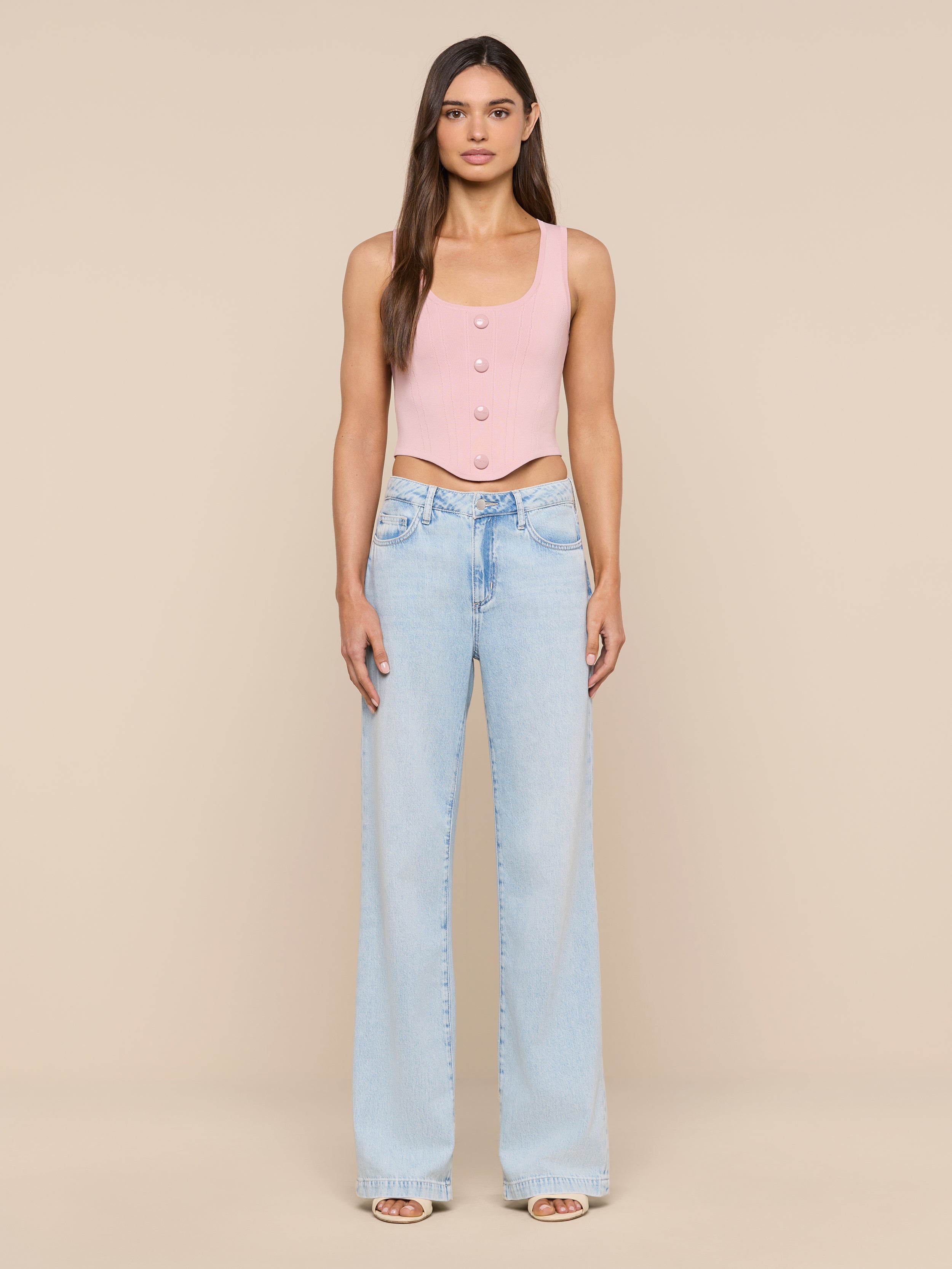 A woman stands facing forward, wearing the Dove Knit Corset Top in pink and light blue wide-leg jeans with a visible waistband detail. The background is plain and white.