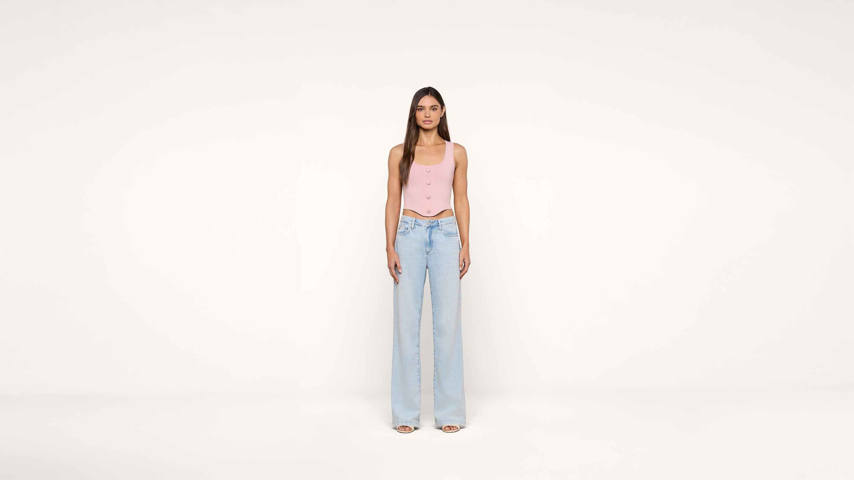 A woman stands facing forward against a plain white background, wearing a light pink sleeveless top and high-waisted, wide-leg light blue jeans with open-toe shoes.