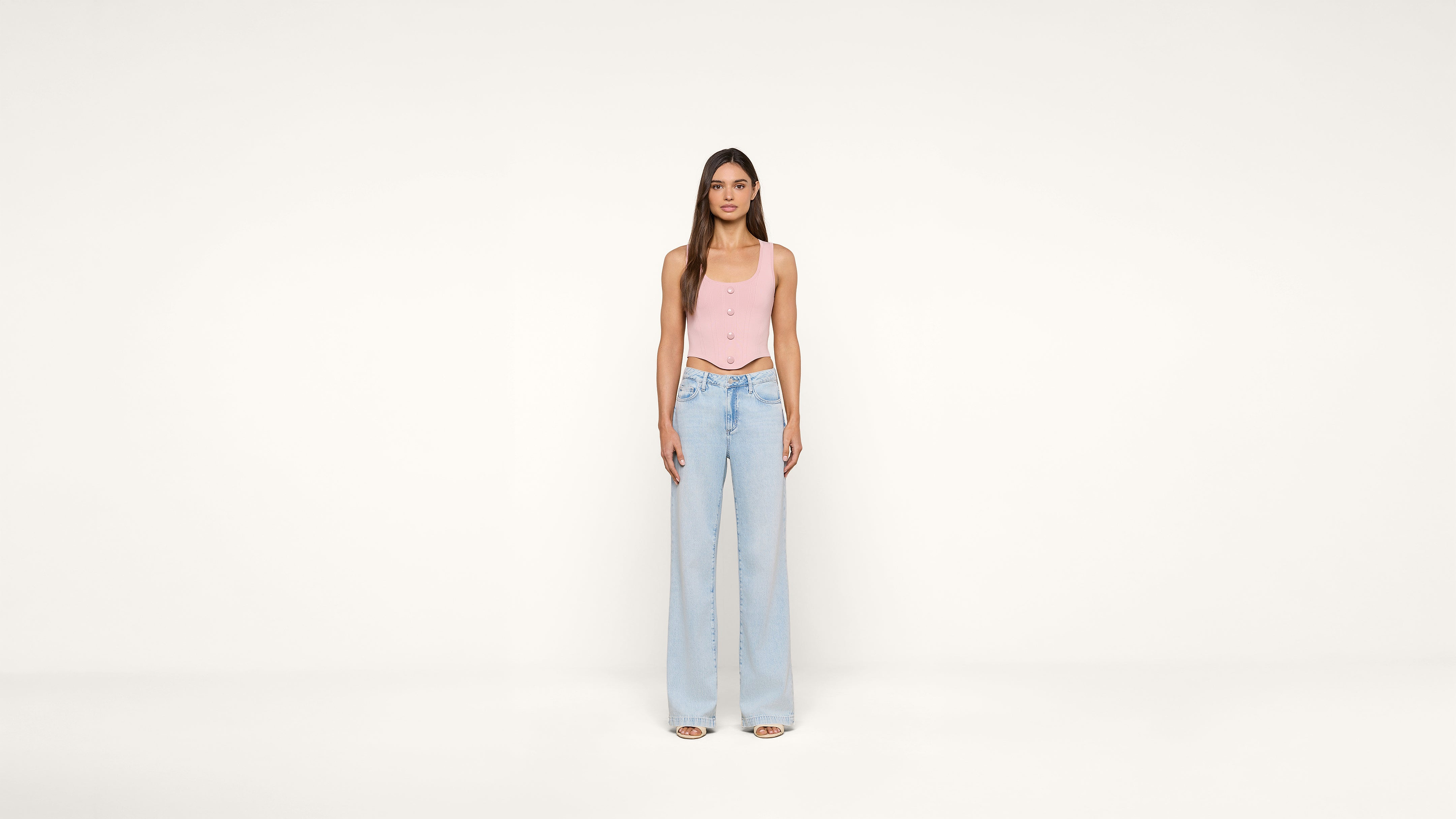 A woman stands facing forward against a plain white background, wearing a light pink sleeveless top and high-waisted, wide-leg light blue jeans with open-toe shoes.