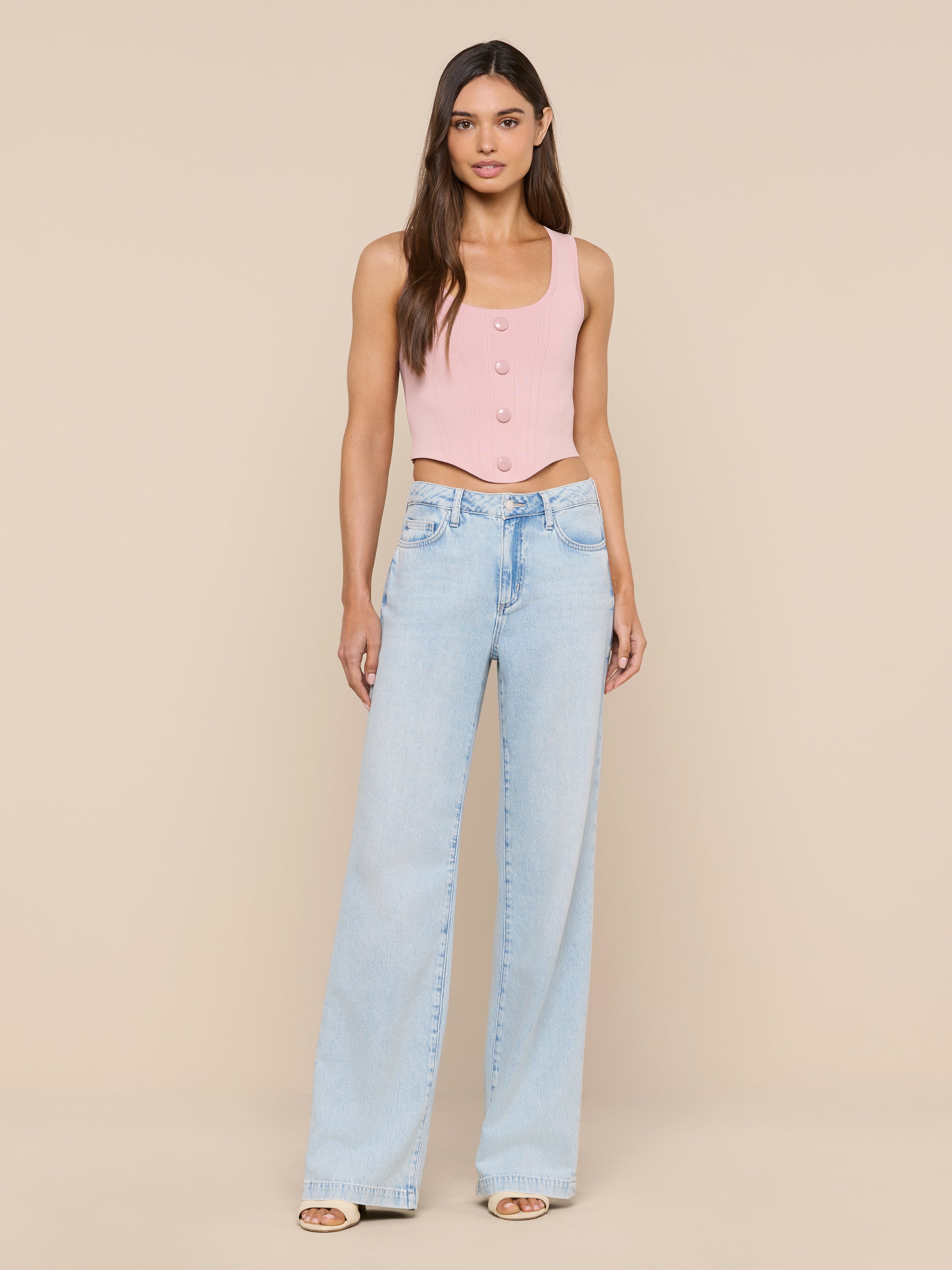 A woman stands facing forward, wearing the Dove Knit Corset Top—a pink fitted sleeveless piece with a curved hemline—paired with light blue wide-leg jeans. She has long, straight hair and is posed against a plain white background.
