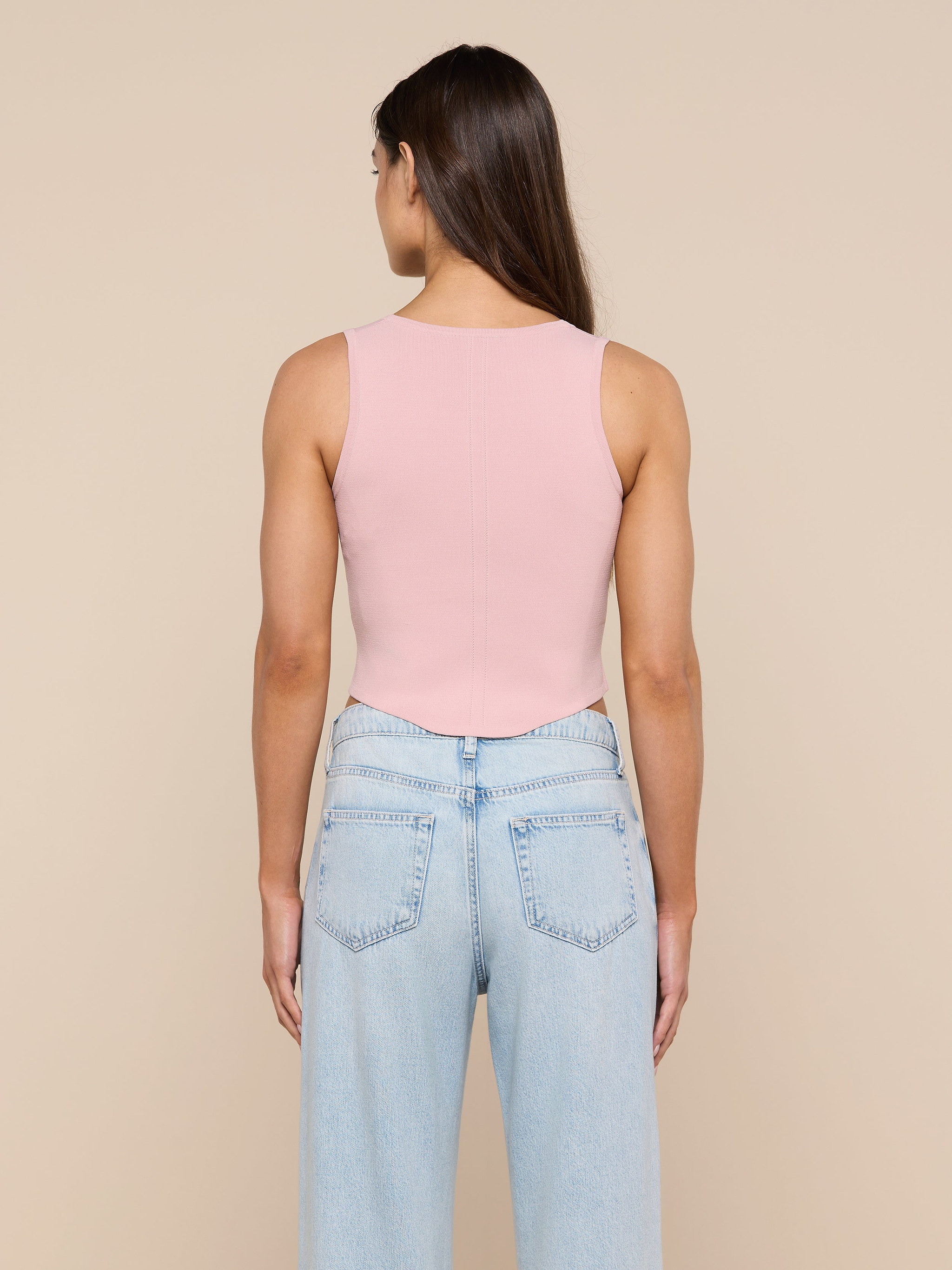 A person with long brown hair, wearing the Dove Knit Corset Top—a sleeveless pink fitted top with a curved hemline—and light blue jeans is standing facing away from the camera against a plain background.