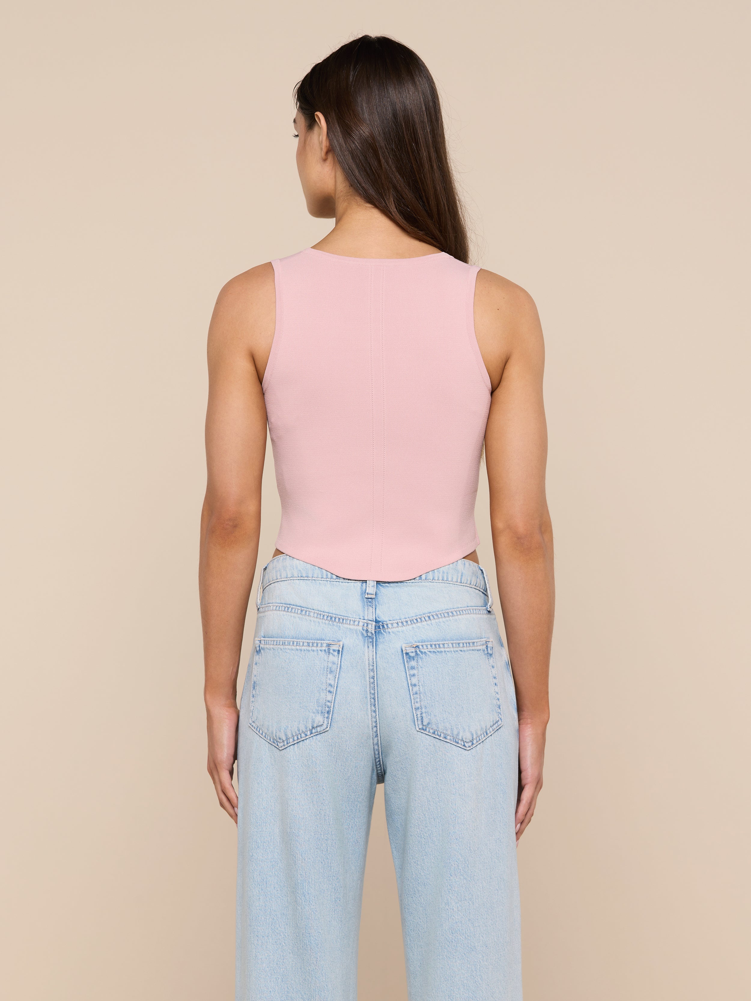 A person with long brown hair, wearing the Dove Knit Corset Top—a sleeveless pink fitted top with a curved hemline—and light blue jeans is standing facing away from the camera against a plain background.