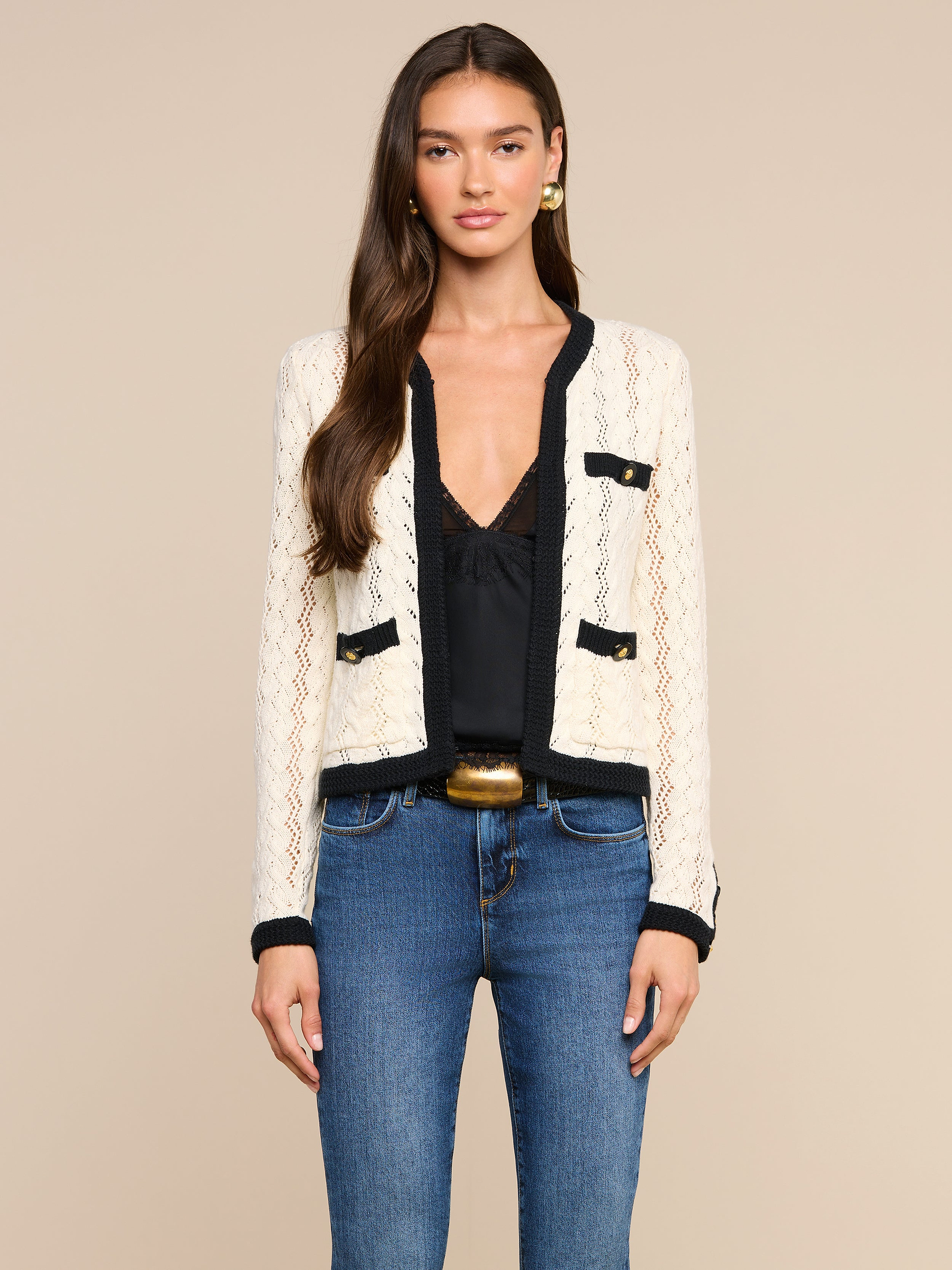 A woman stands facing forward against a plain light background, wearing the Zephyra Pointelle Knit Cardigan—a cream knit with black trim and decorative buttons—layered over a black top and blue jeans.