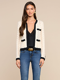 A woman stands facing forward against a plain light background, wearing the Zephyra Pointelle Knit Cardigan—a cream knit with black trim and decorative buttons—layered over a black top and blue jeans.