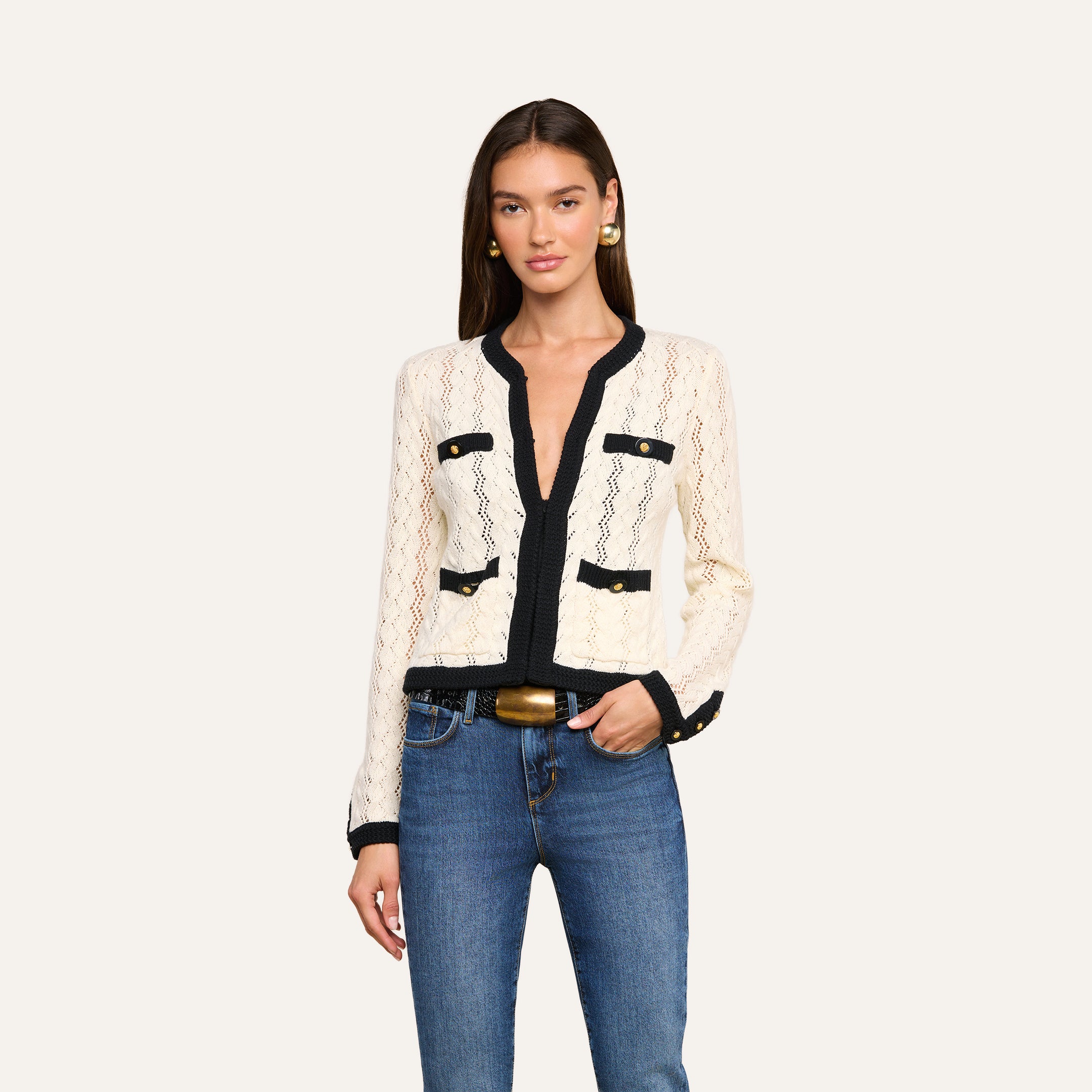 A woman wearing the Zephyra Pointelle Knit Cardigan in cream with black trim and decorative gold buttons, styled with blue jeans and gold earrings, stands against a plain light background.