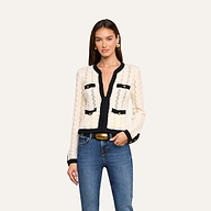 A woman wearing the Zephyra Pointelle Knit Cardigan in cream with black trim and decorative gold buttons, styled with blue jeans and gold earrings, stands against a plain light background.