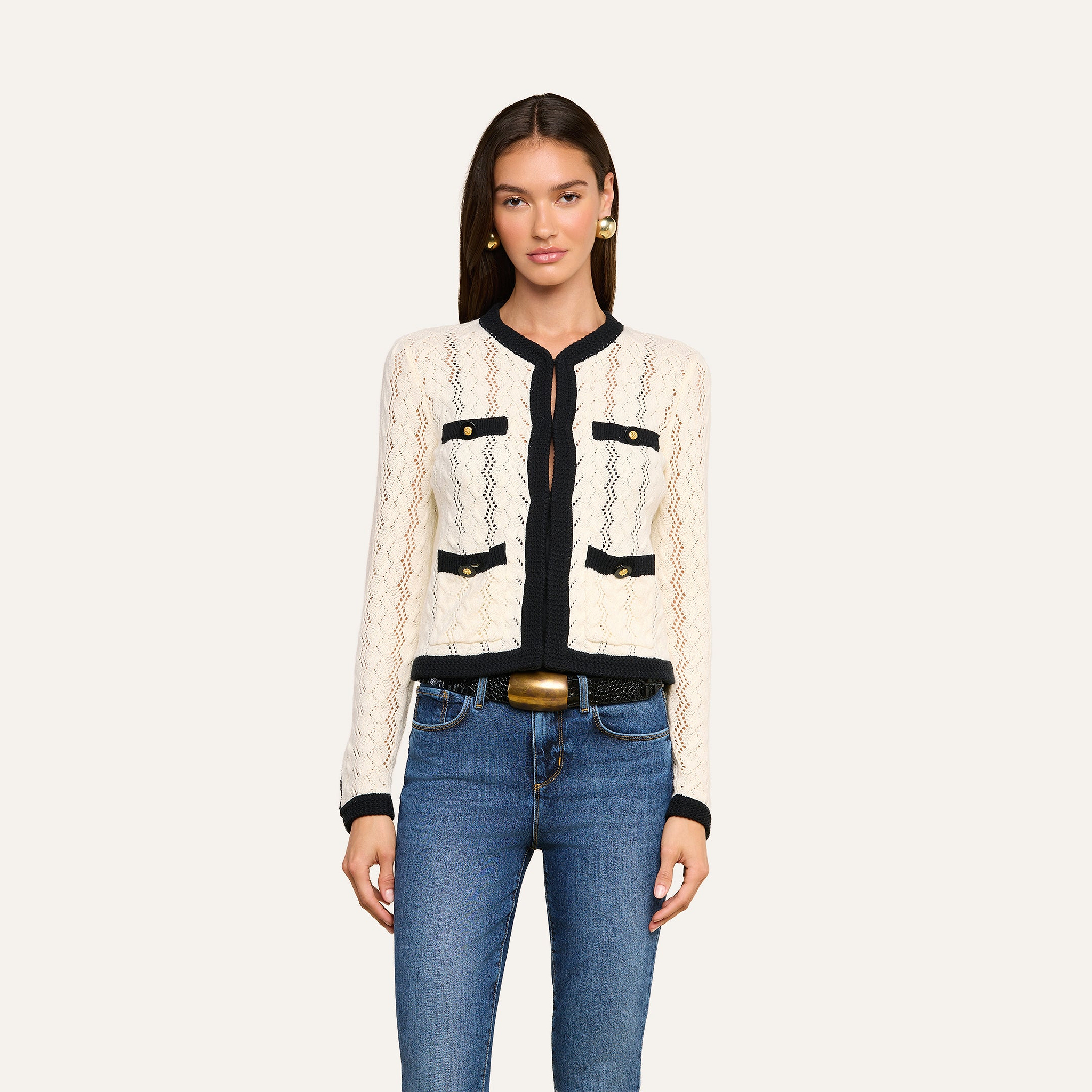 A woman poses against a plain light background wearing the Zephyra Pointelle Knit Cardigan in cream with black trim and decorative gold buttons, styled with blue jeans, a belt, and gold earrings.