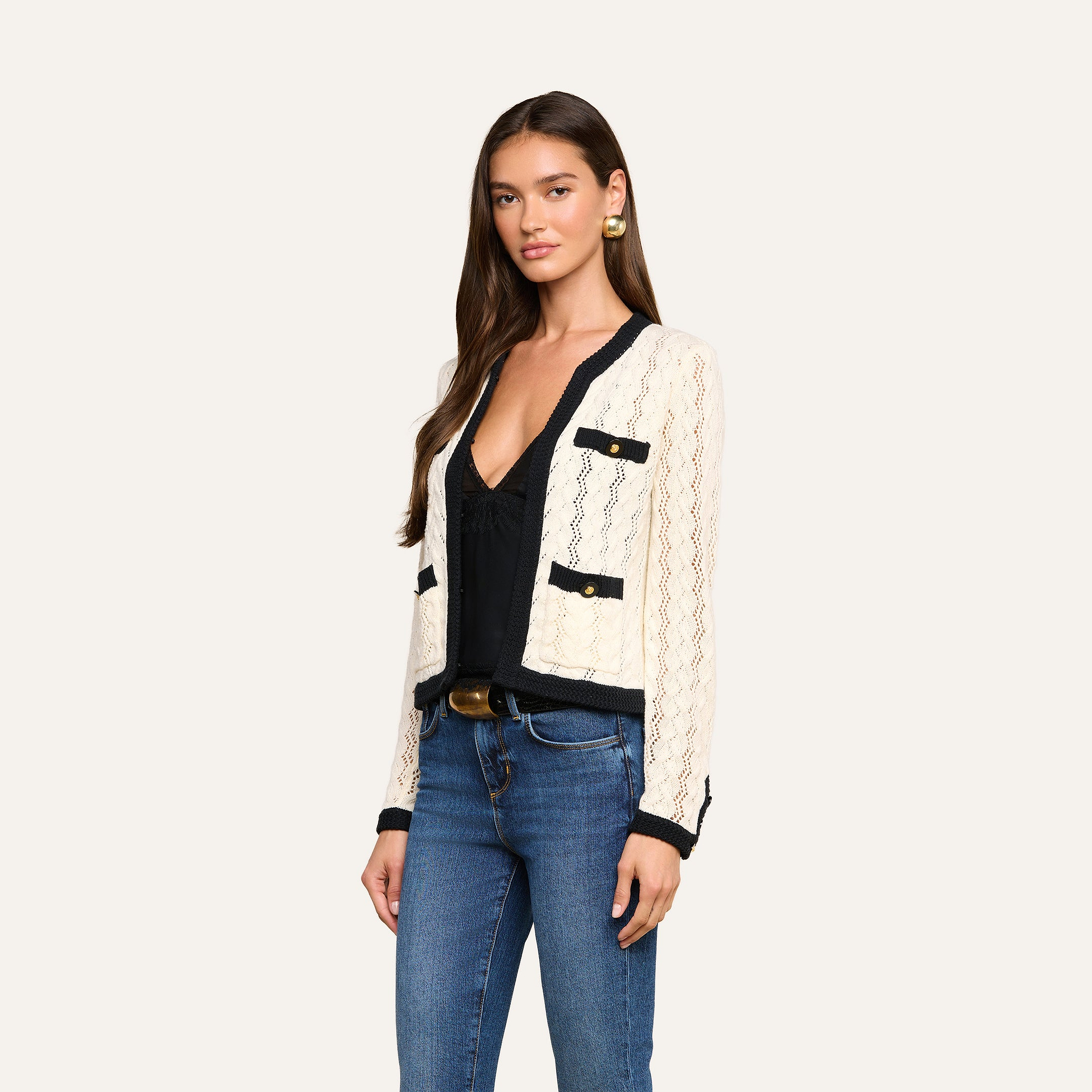 A woman wearing the cream Zephyra Pointelle Knit Cardigan with black trim and decorative buttons, paired with a black top, blue jeans, and gold earrings, stands against a plain light background.