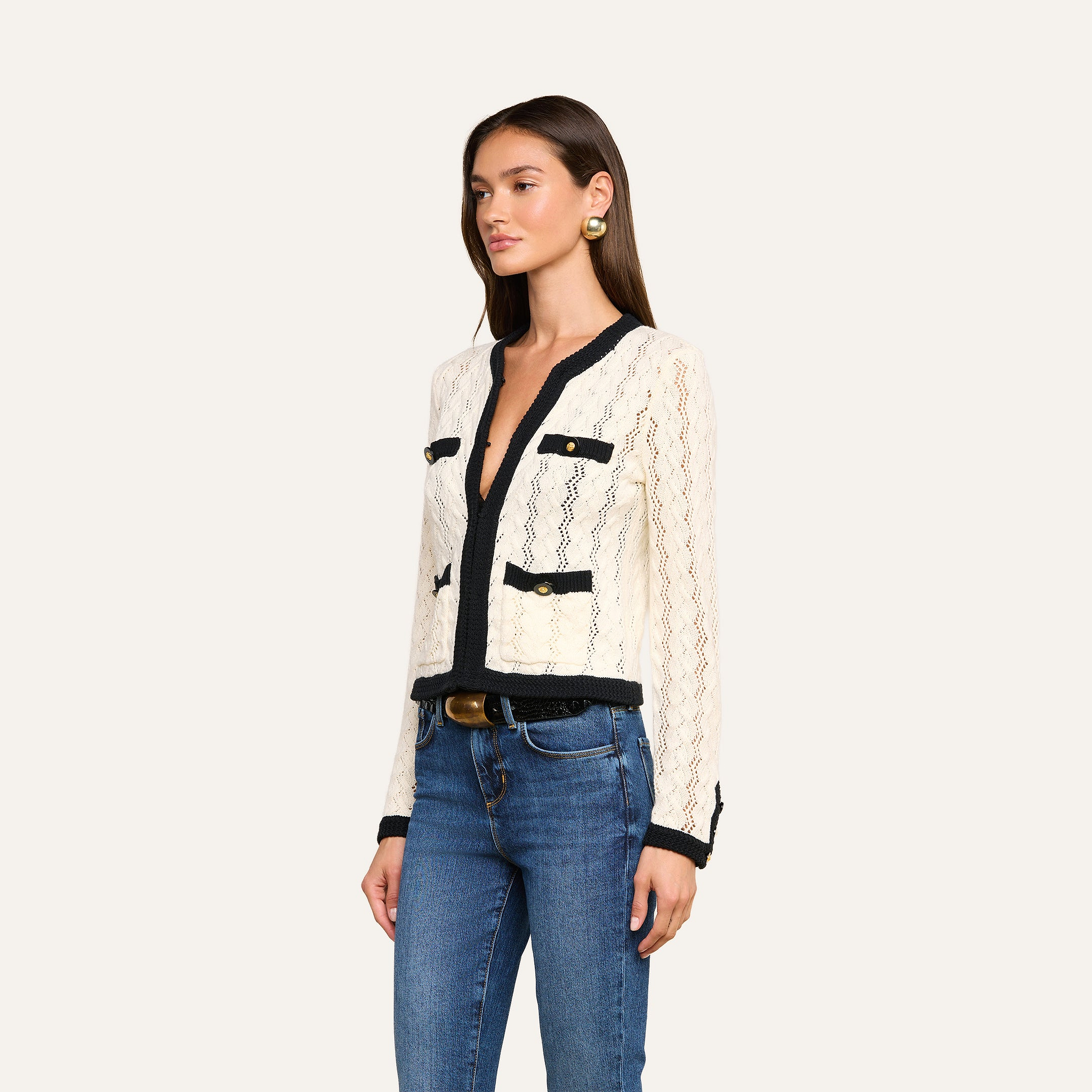 A woman wears the Zephyra Pointelle Knit Cardigan in cream with black trim, featuring long sleeves, decorative buttons, and pockets, styled with blue jeans. She stands against a plain light background, angled slightly to the side.