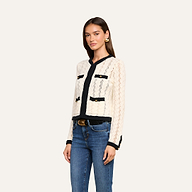 A woman with long brown hair wears the Zephyra Pointelle Knit Cardigan, featuring a cream color with black trim and decorative gold buttons. She pairs it with blue jeans, a black belt, and large gold earrings. She stands against a plain light background, facing slightly to the right.
