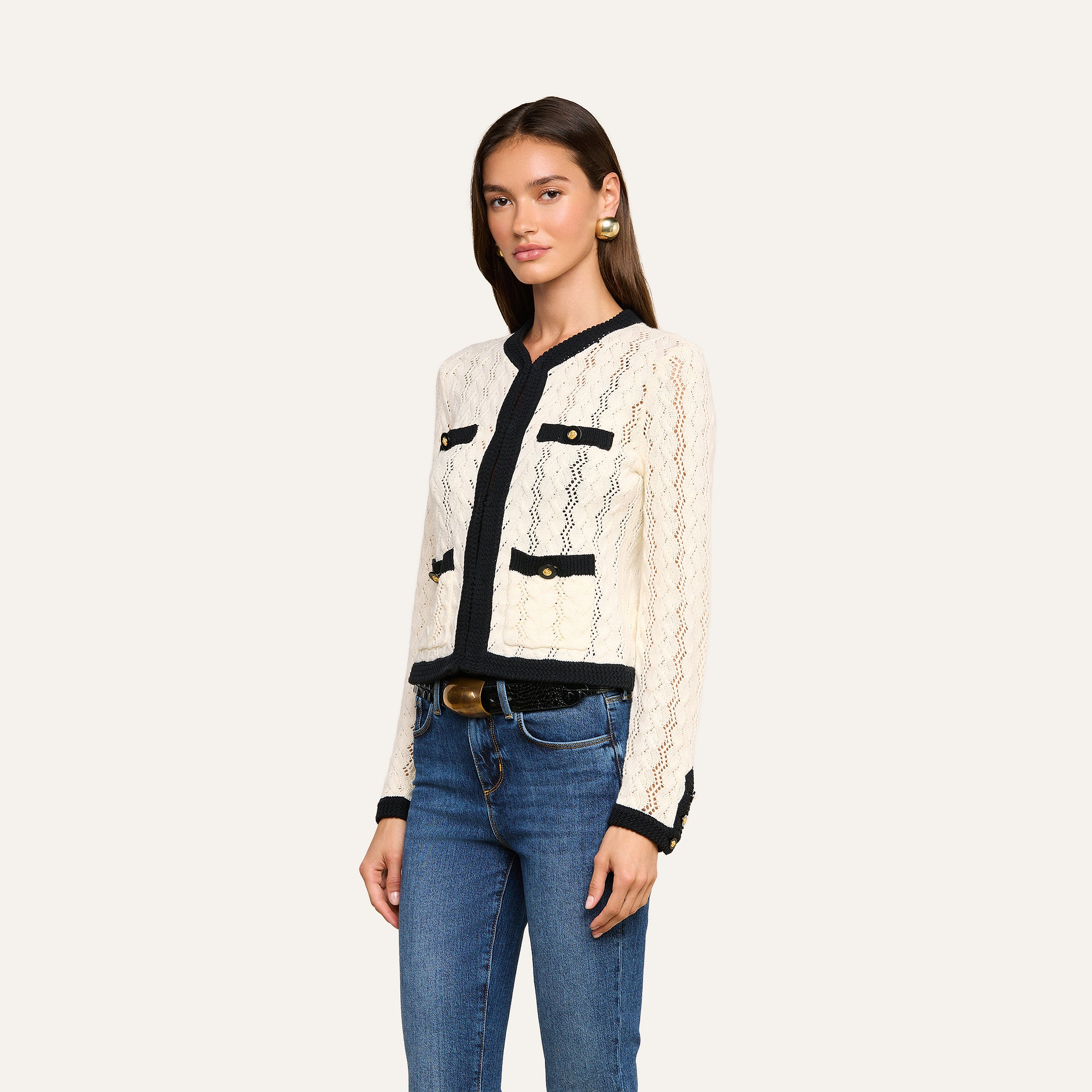 A woman with long brown hair wears the Zephyra Pointelle Knit Cardigan, featuring a cream color with black trim and decorative gold buttons. She pairs it with blue jeans, a black belt, and large gold earrings. She stands against a plain light background, facing slightly to the right.