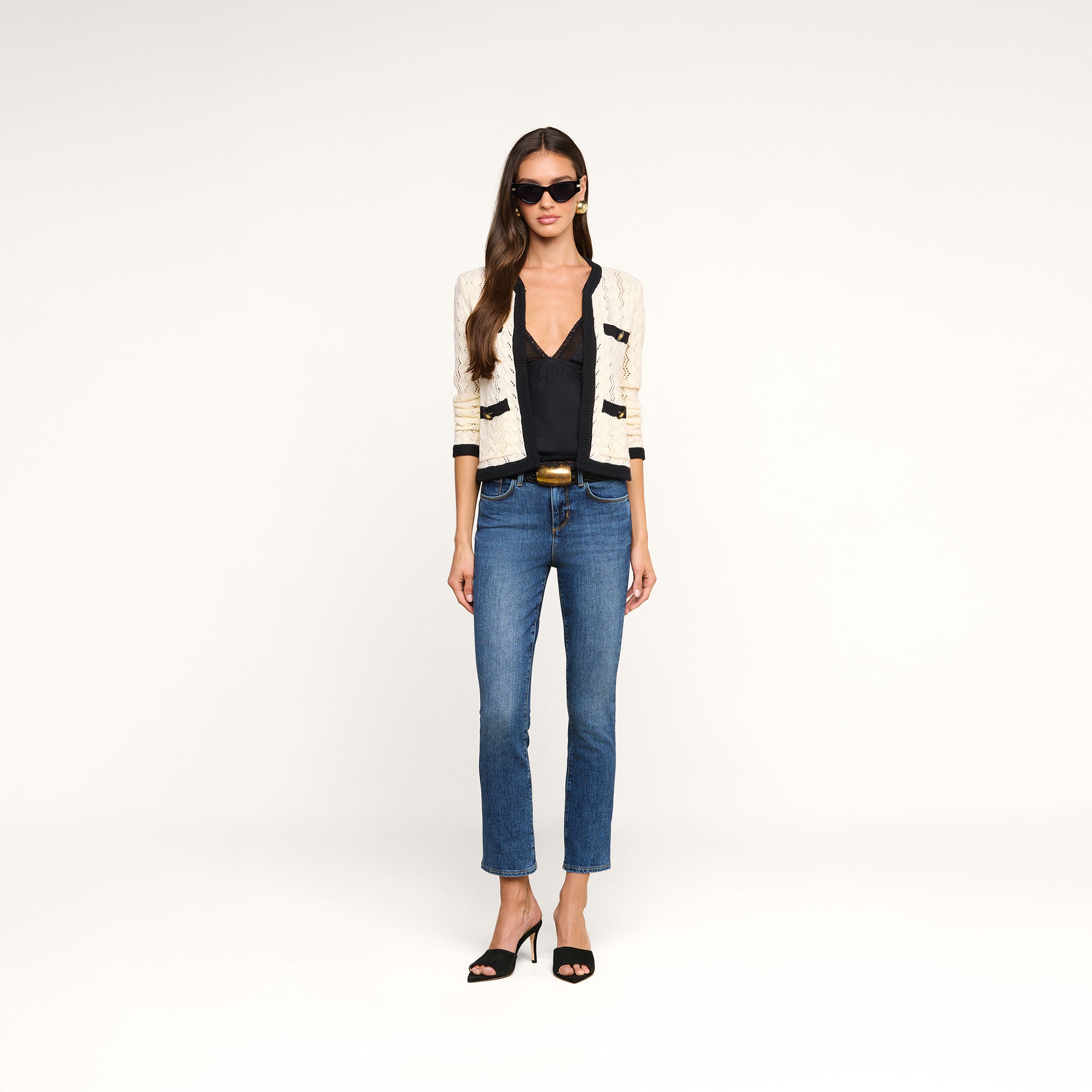 A woman wearing sunglasses, a cream-colored Zephyra Pointelle Knit Cardigan with decorative buttons, a black top, blue jeans, and black heeled sandals stands against a plain white background.