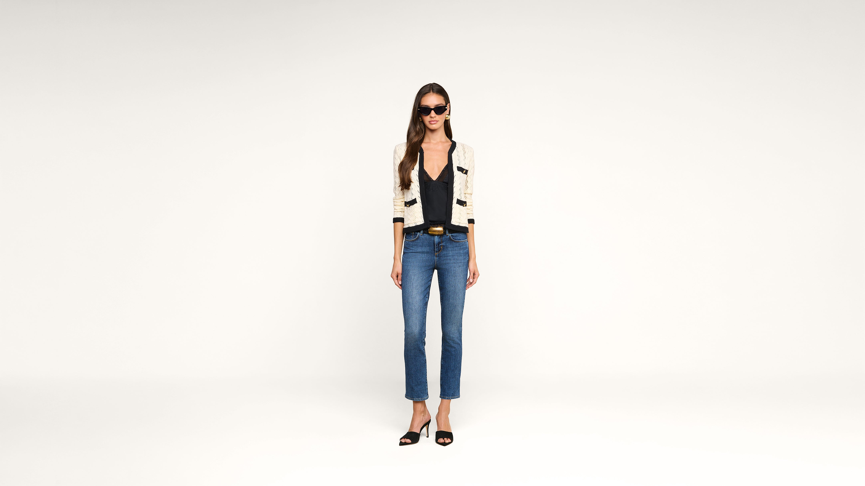 Woman in sunglasses wearing a cream cardigan with black trim, black top, blue jeans, and black heels stands against a plain white background.
