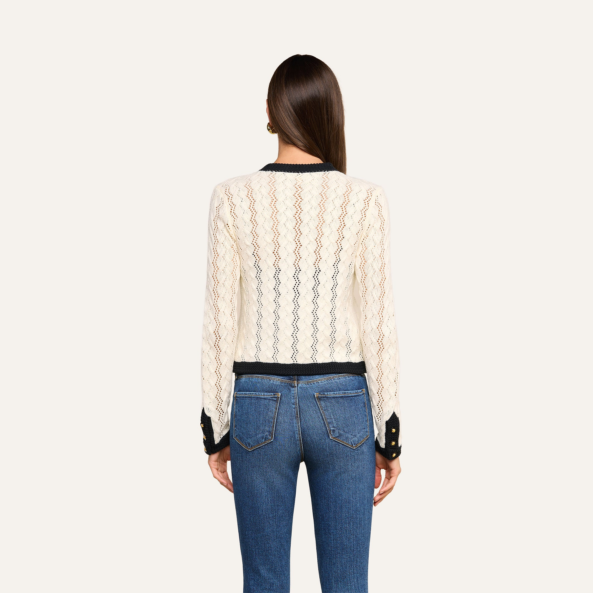 A woman with straight brown hair, wearing the Zephyra Pointelle Knit Cardigan in cream with black trim and blue jeans, stands facing away from the camera against a plain light background.
