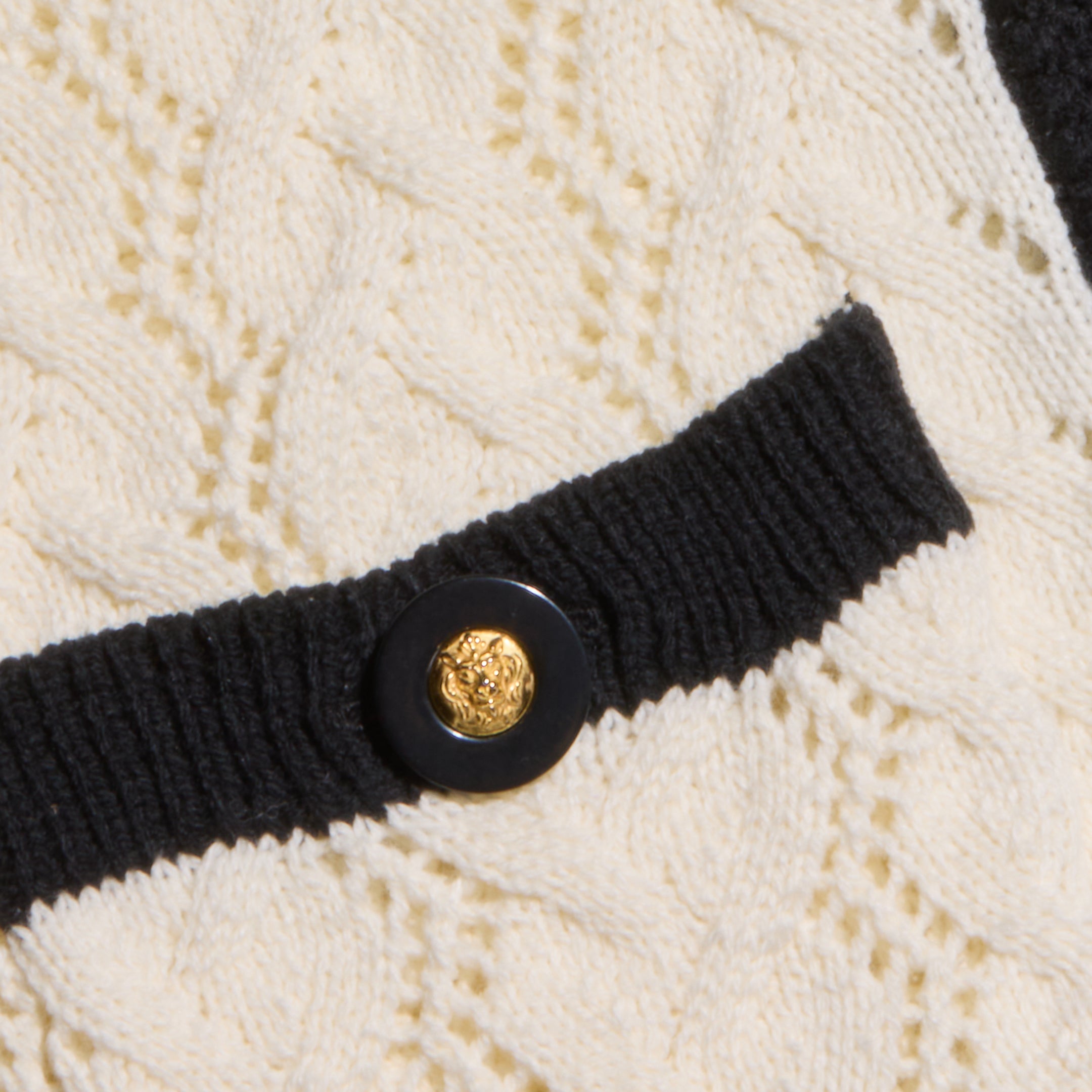 Close-up of the Zephyra Pointelle Knit Cardigan in cream, featuring a textured pointelle knit with a black ribbed edge and decorative buttons adorned with gold center details.