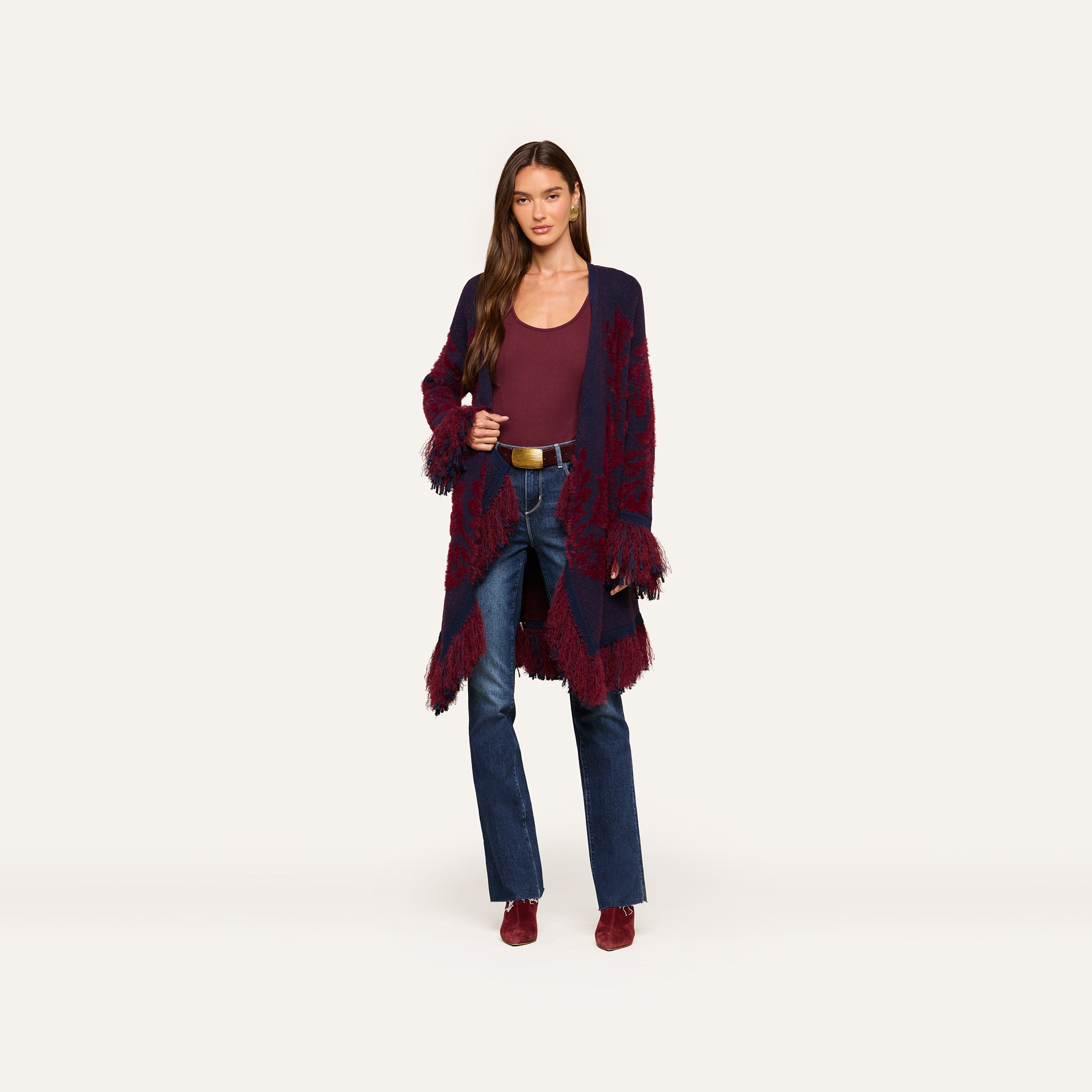 A woman stands against a plain white background wearing a burgundy top, blue jeans, maroon shoes, and the Naiad Jacquard Fringe Cardigan—a long boho chic piece with vintage fringe edges in maroon and navy jacquard knit.