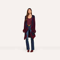 A woman stands against a plain light background, wearing a maroon top, blue jeans, maroon shoes, a brown belt, and the Naiad Jacquard Fringe Cardigan in maroon and navy.
