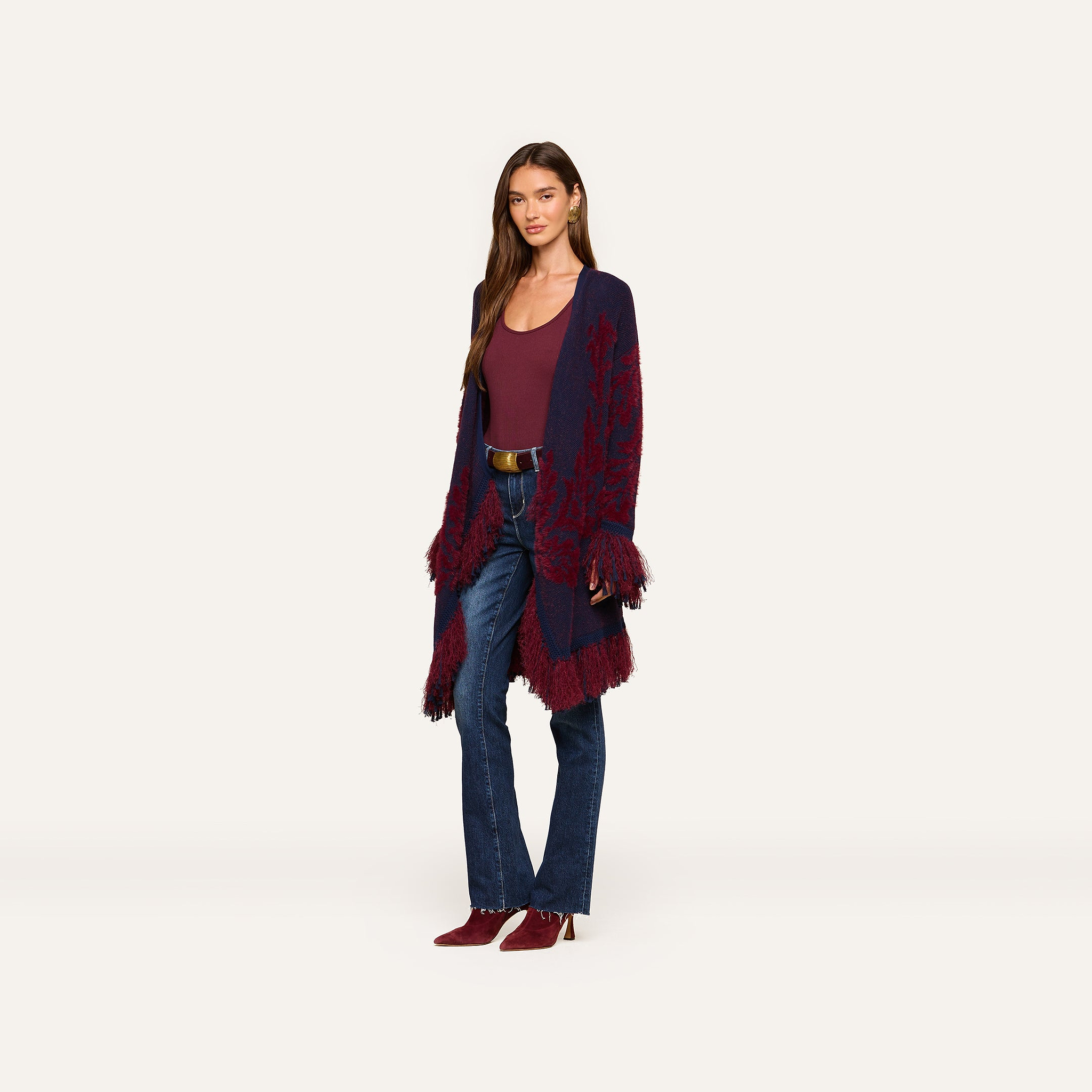 A woman stands against an off-white background wearing the Naiad Jacquard Fringe Cardigan in dark purple and red, styled with a maroon top, blue jeans, and maroon shoes.