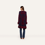 A woman stands facing away, wearing the Naiad Jacquard Fringe Cardigan featuring boho chic navy and burgundy patterns with vintage-inspired fringe at the hem and sleeves, styled with blue jeans and heeled shoes against a plain light background.
