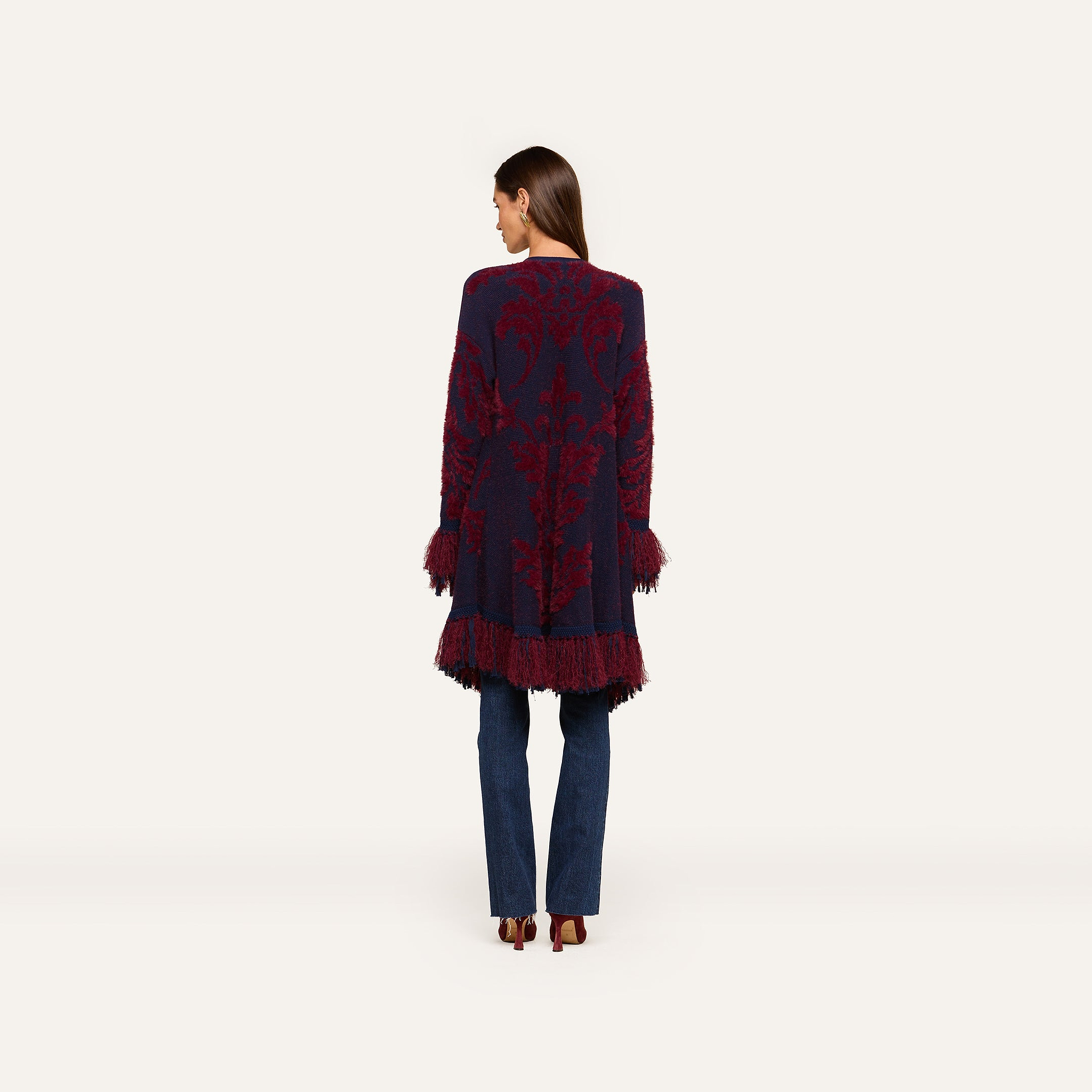 A woman stands facing away, wearing the Naiad Jacquard Fringe Cardigan featuring boho chic navy and burgundy patterns with vintage-inspired fringe at the hem and sleeves, styled with blue jeans and heeled shoes against a plain light background.