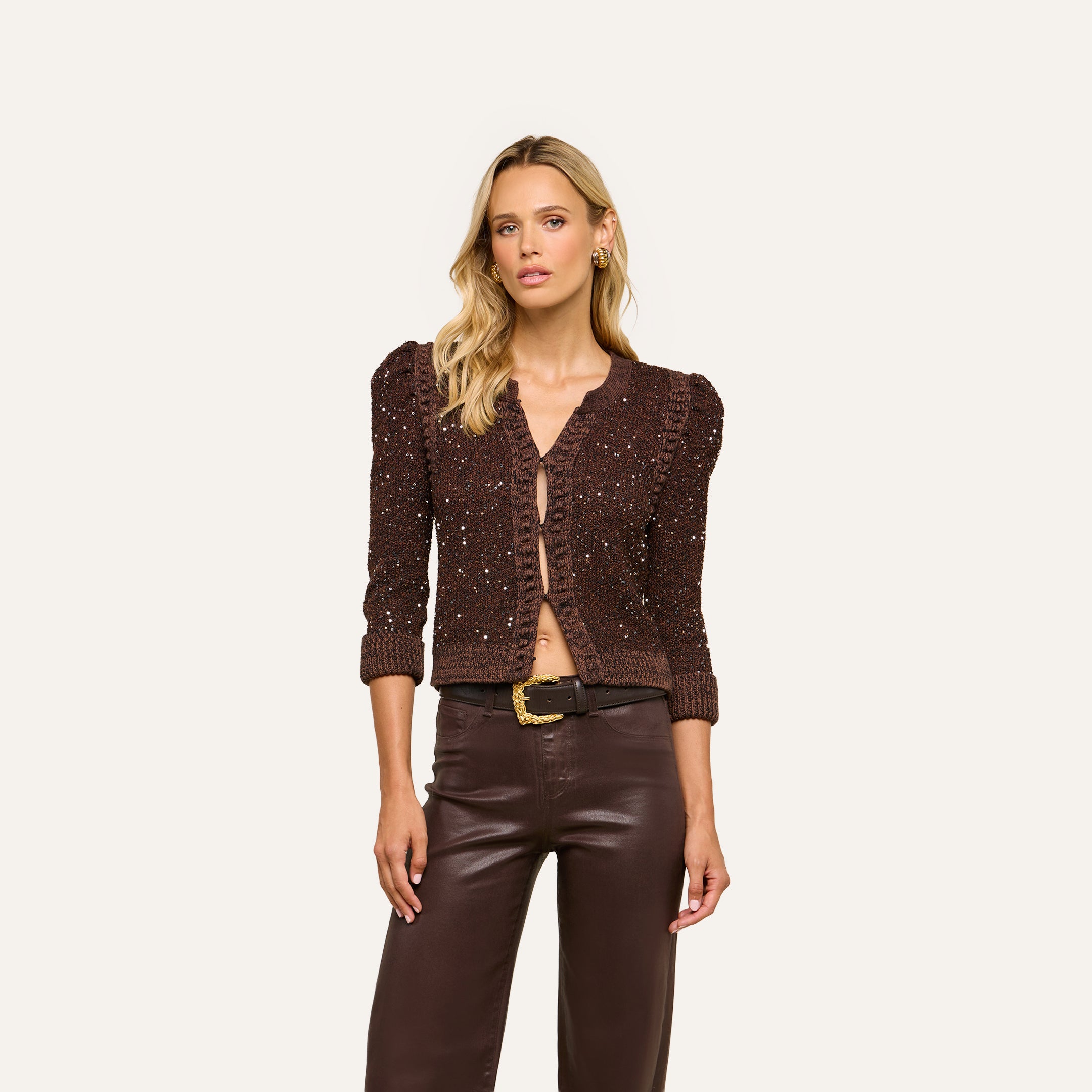 Wearing a Thistle Sequined Cardigan with puffed sleeves and brown leather pants, this woman exudes chic New York Fashion Week style against a simple light background.