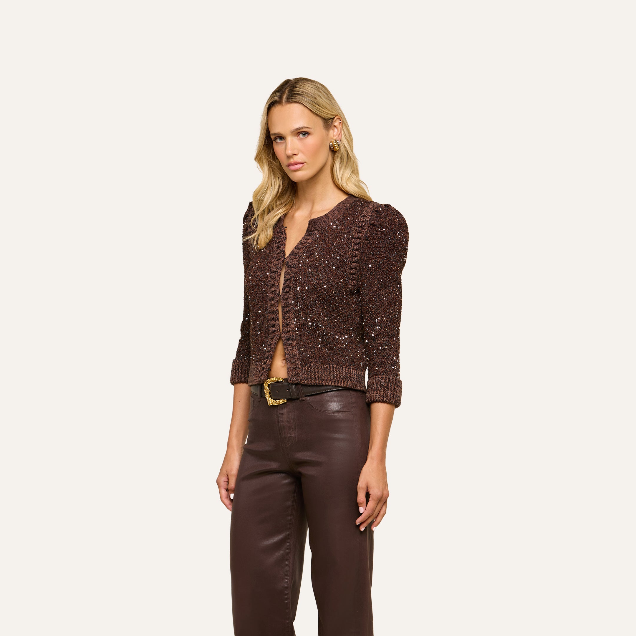 Wearing the Thistle Sequined Cardigan and brown leather pants, a woman stands against a plain light background, gazing slightly to the side with a neutral expression.