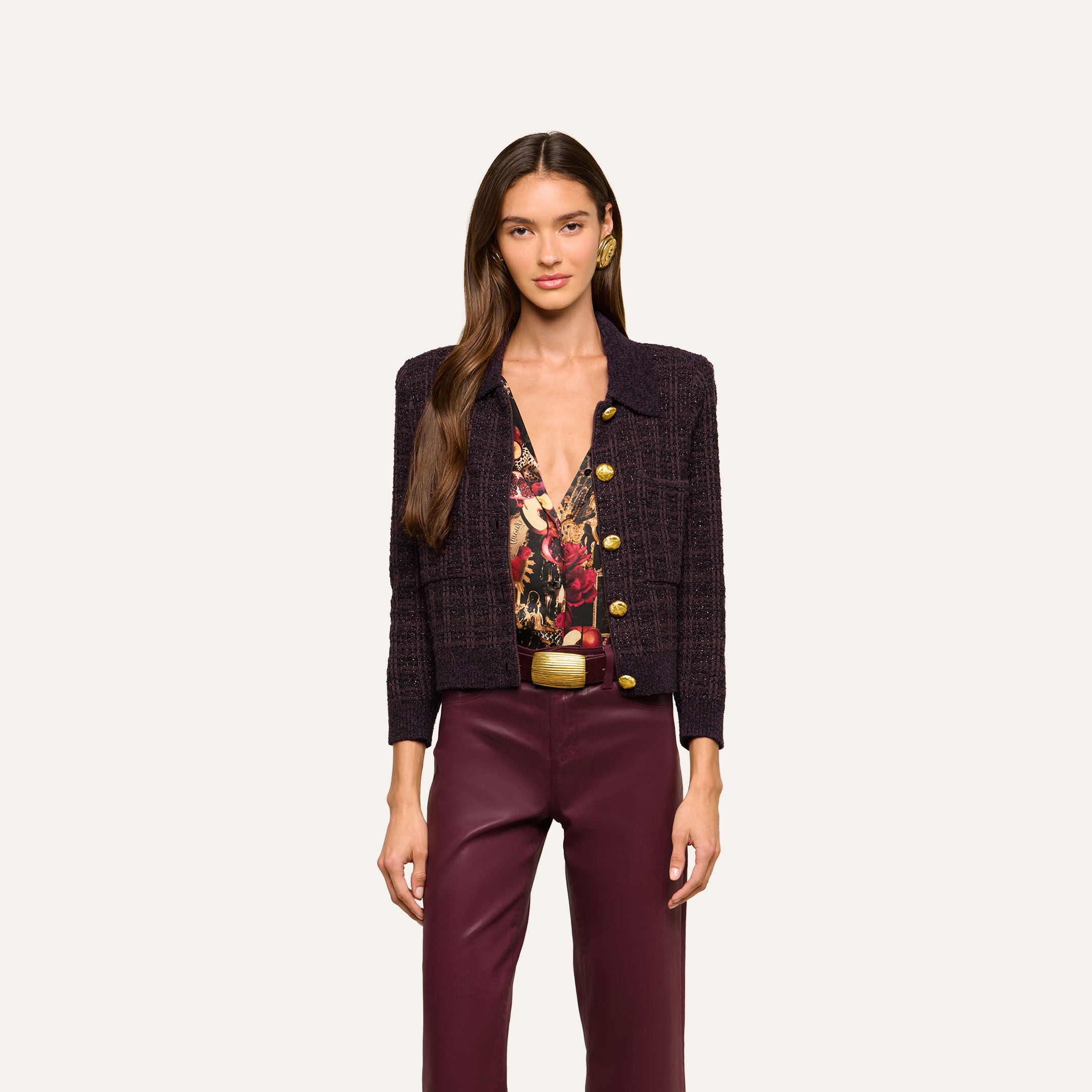 A woman stands facing forward against a plain light background, wearing the Morrigan Cardigan in dark brown, paired with a patterned blouse and burgundy pants.