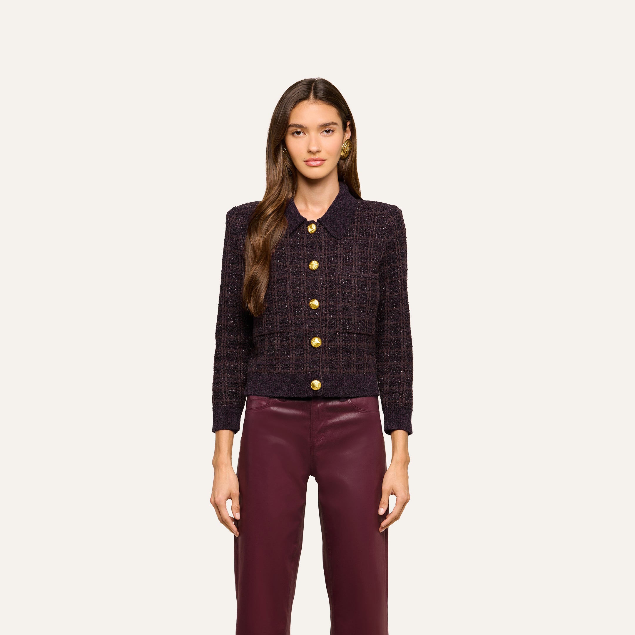 A woman with long brown hair wears the Morrigan Cardigan—a dark brown tweed piece with gold buttons—paired with burgundy pants, standing against a plain light background.