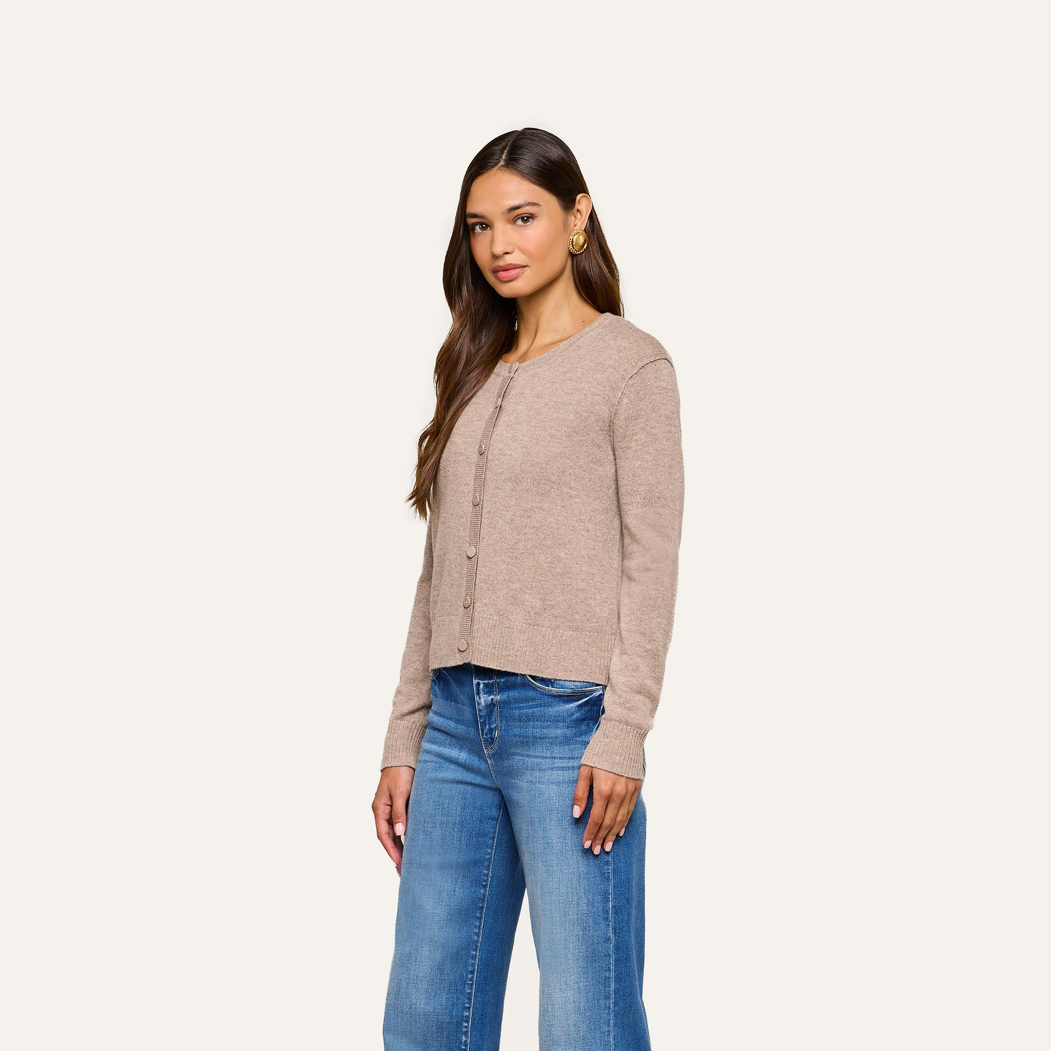 A woman wearing the Malone Cashmere Cardigan and blue jeans stands against a plain off-white background, looking slightly to the side with a neutral expression.