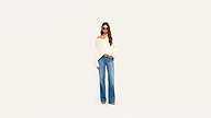 Woman in sunglasses, off-shoulder white sweater, and wide-leg blue jeans stands confidently against a plain white background.