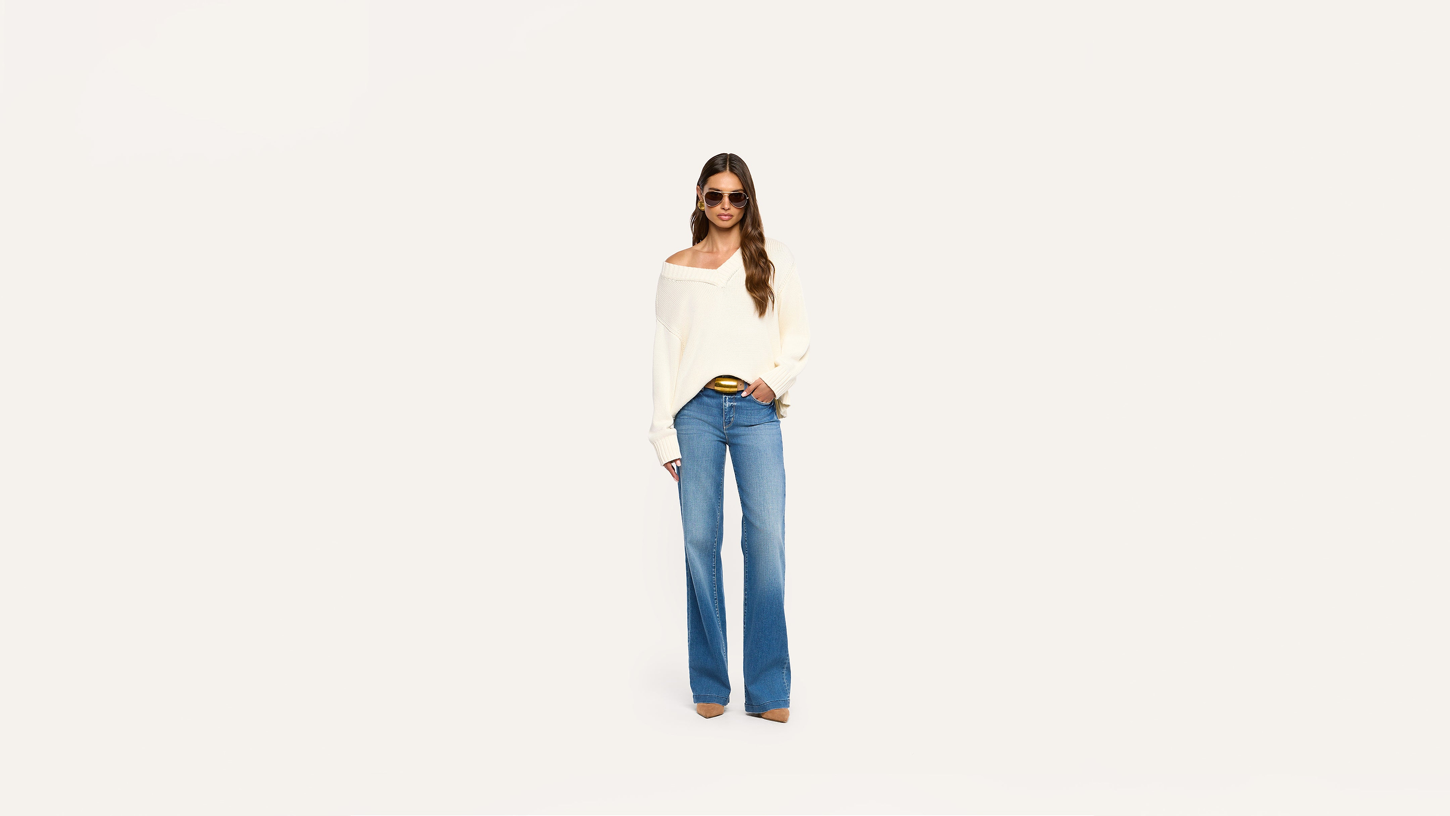 Woman in sunglasses, off-shoulder white sweater, and wide-leg blue jeans stands confidently against a plain white background.