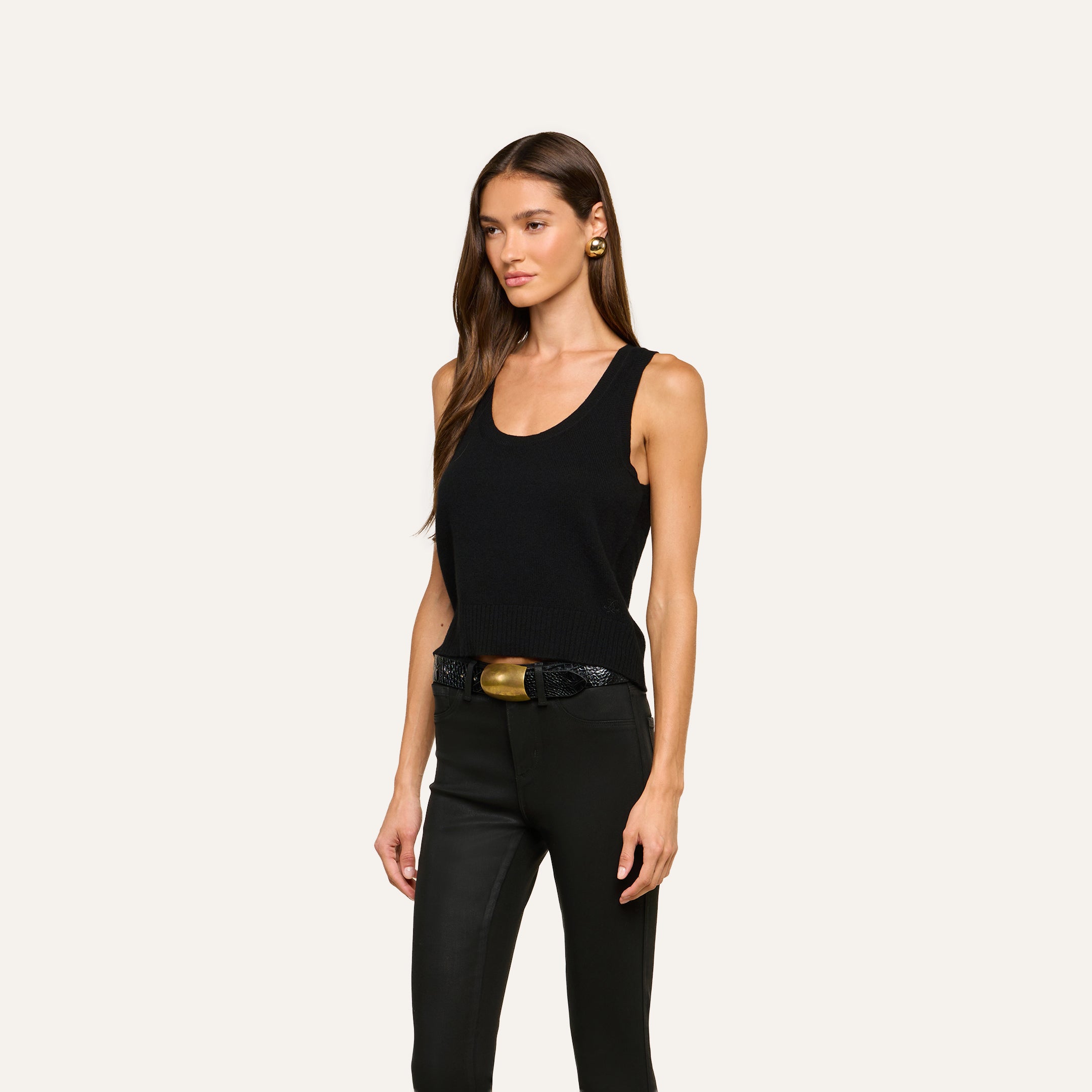 A woman with long brown hair wears the Filou Cashmere Tank—a black, sleeveless top—paired with black pants, a black belt featuring a gold buckle, and gold earrings for an effortlessly luxurious layered look. She stands against a plain light background.