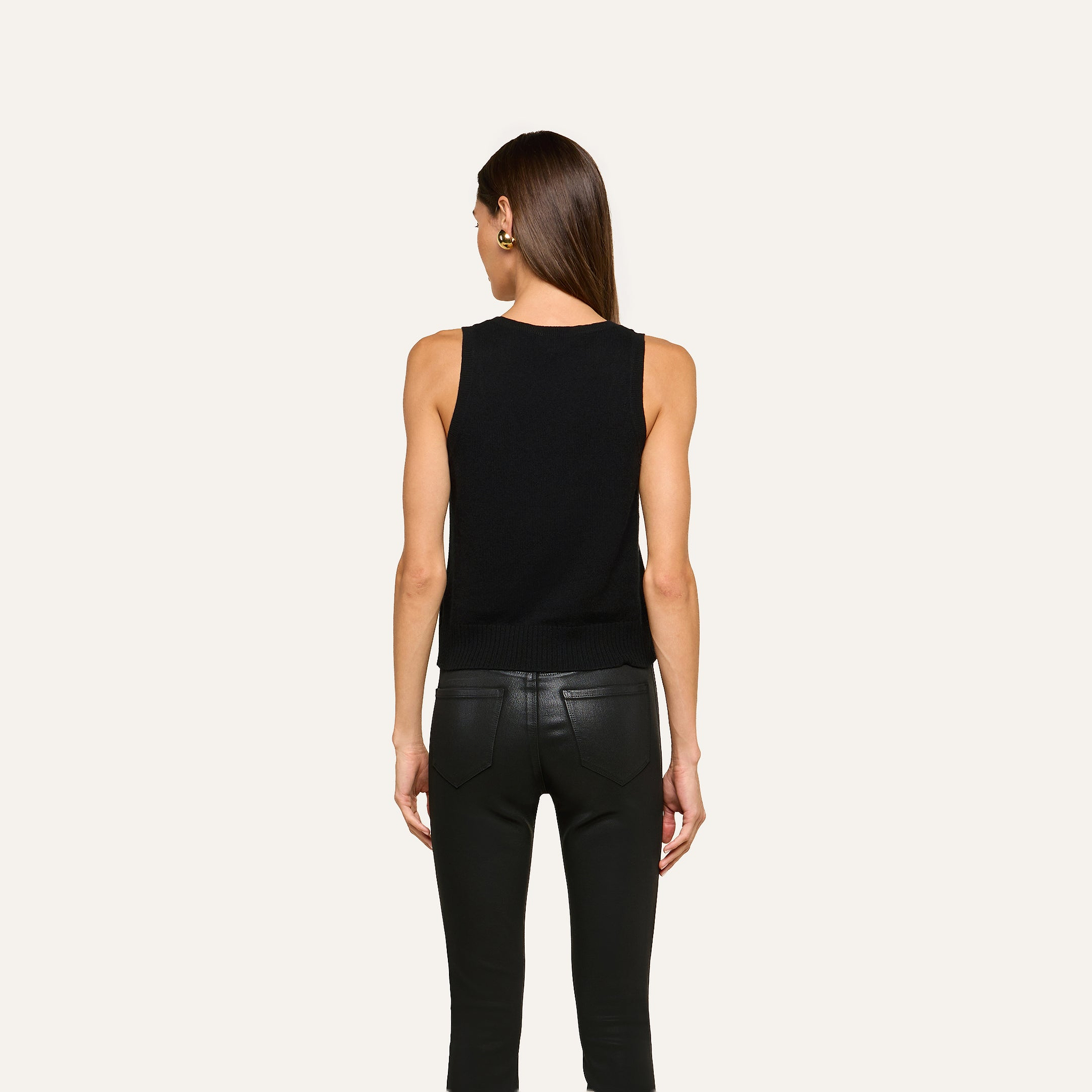 A woman with straight brown hair, wearing the Filou Cashmere Tank in black and black leather pants, stands facing away against a plain light background.