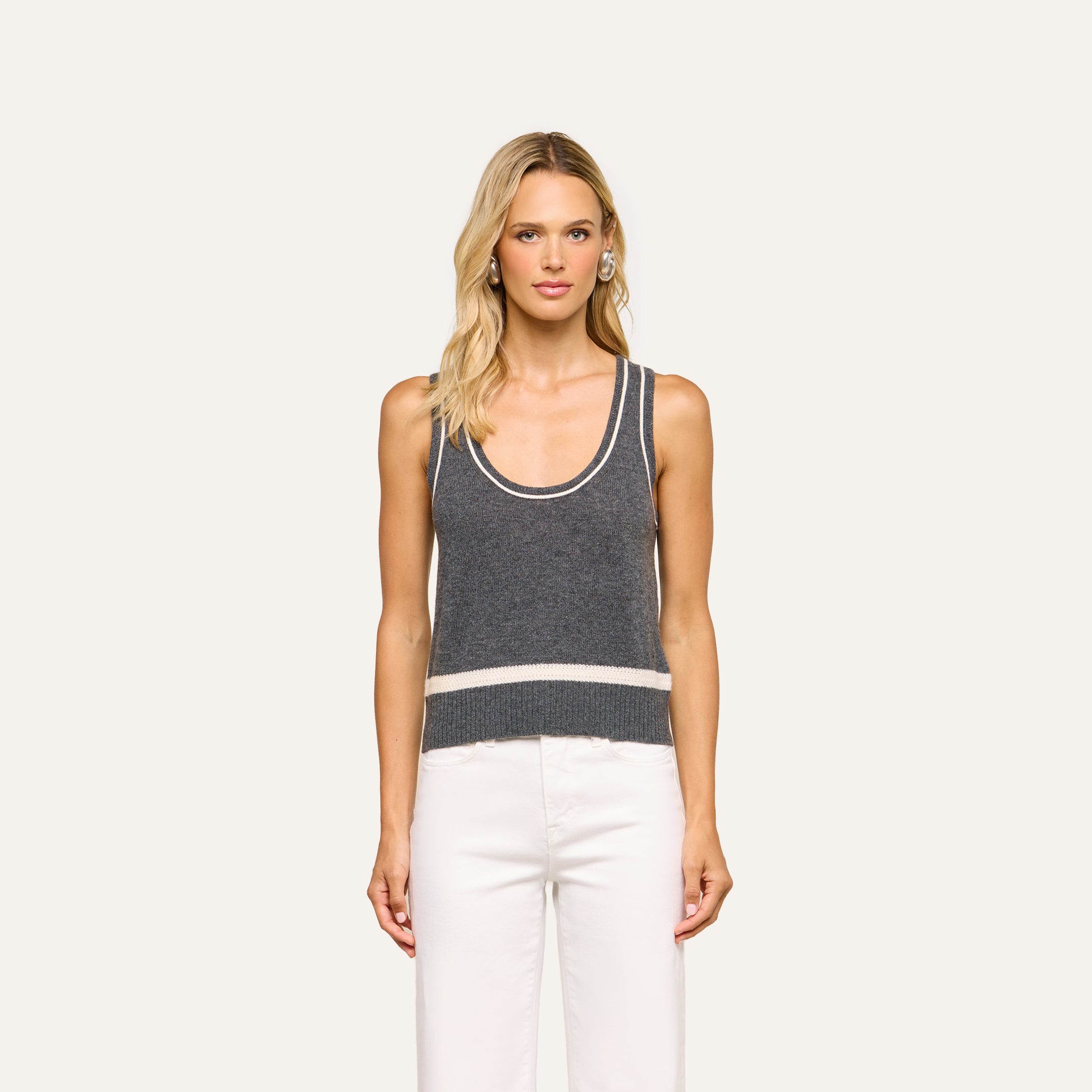 A woman with long blonde hair stands facing forward against a plain light background, wearing the Filou Cashmere Tank—a sleeveless gray knit top with white trim—paired with white pants.