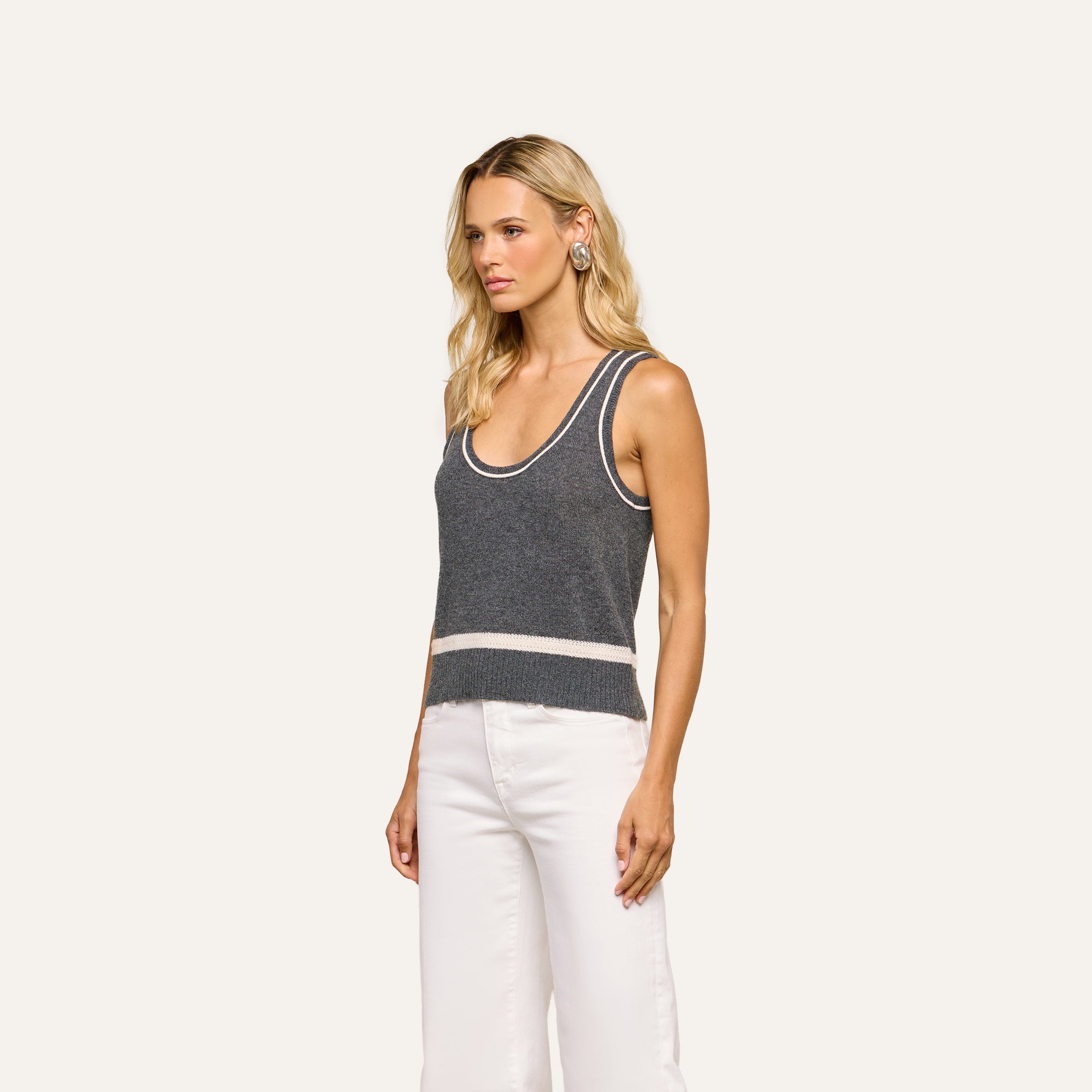 Wearing the Filou Cashmere Tank—a gray lightweight cashmere sleeveless knit top with white trim—a woman stands against a plain light background in white pants, looking to the side.