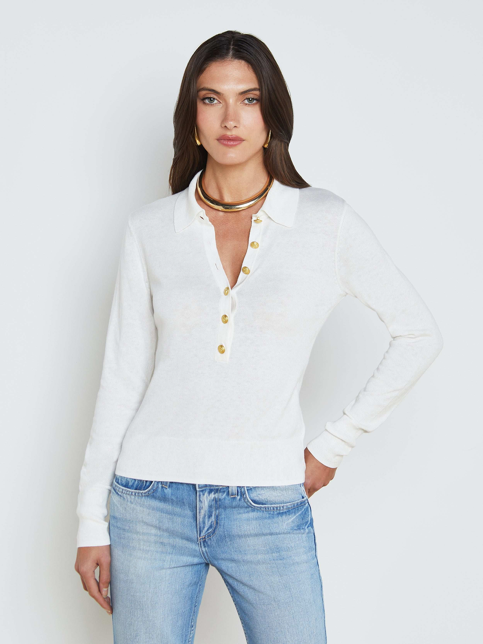 A woman models the Sterling Sweater, a slim fit white long sleeve knit shirt, paired with blue jeans as she stands with one hand on her hip against a plain light background.