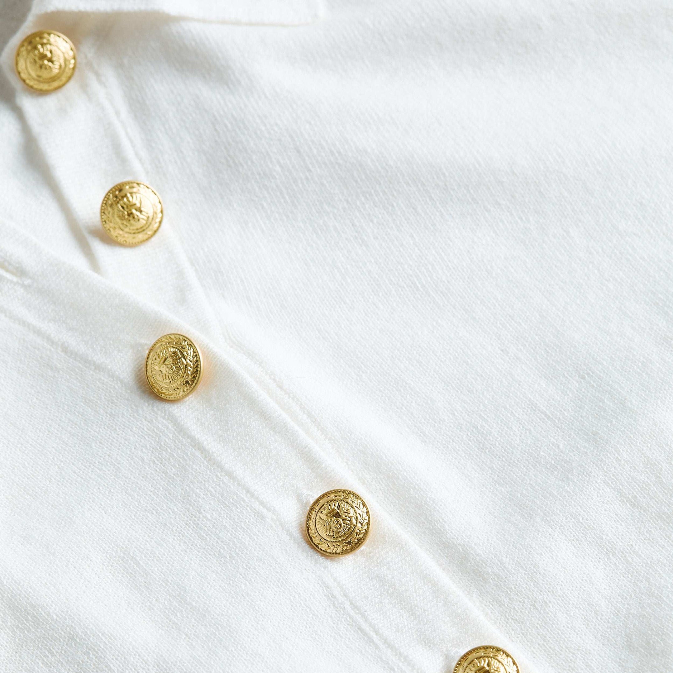 Close-up of the Sterling Sweater’s premium white knit fabric adorned with five shiny gold buttons in a row, each featuring an embossed design.