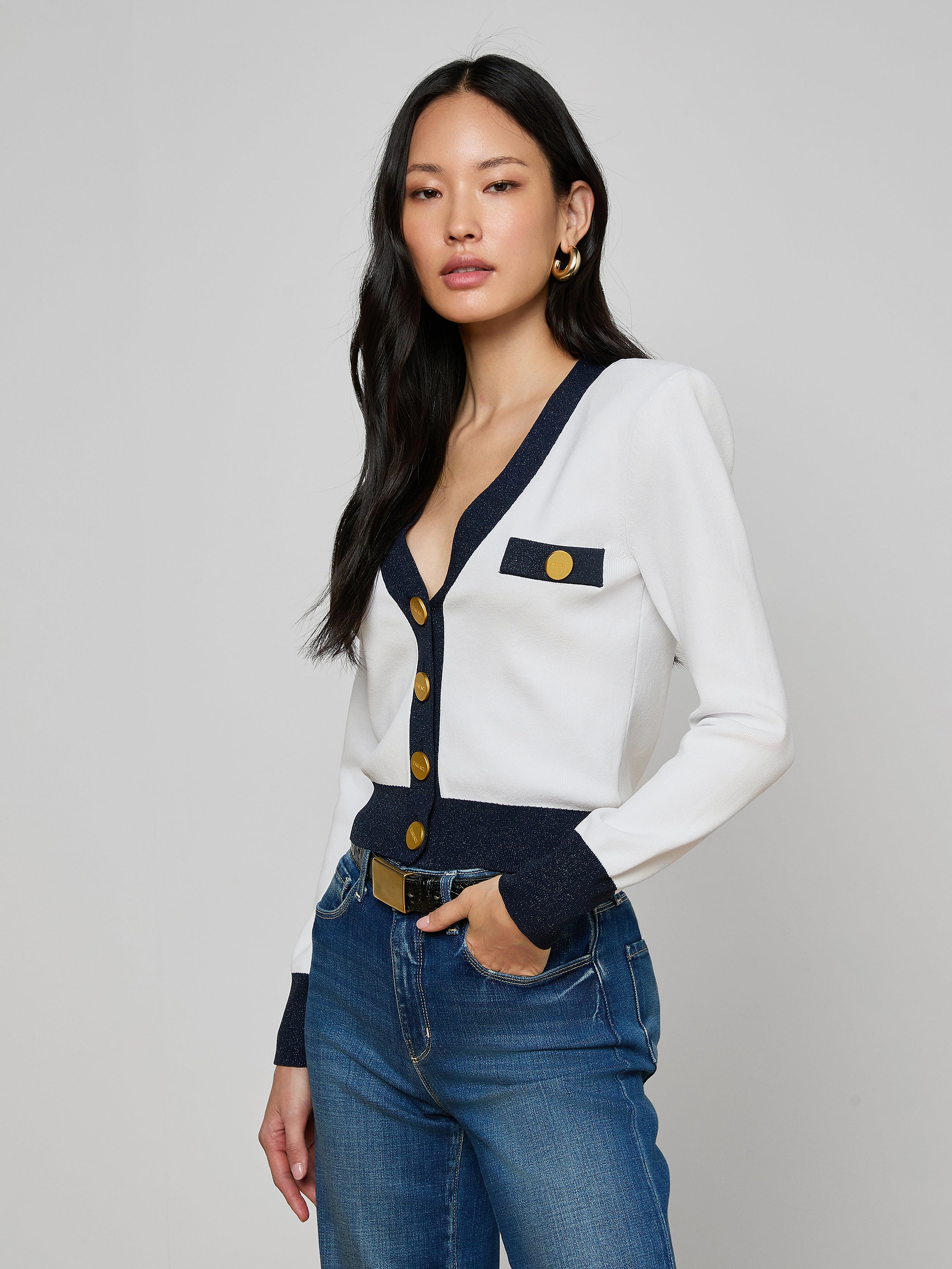 A woman with long black hair wears the Taya Cardigan, a Parisian-inspired white piece featuring navy and metallic trim with gold buttons, paired with blue jeans. She stands with one hand in her pocket against a plain light background.