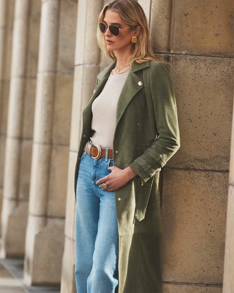 Una donna che indossa occhiali da sole, un lungo cappotto verde oliva, un top color crema, jeans blu e una cintura marrone sta in piedi contro un pilastro di pietra, guardando di lato con una mano in tasca.