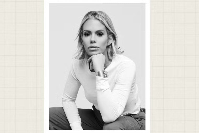 Black and white photo of a woman with light hair wearing a long-sleeve shirt, resting her chin on her hand and looking at the camera with a neutral expression. She is seated against a plain background.